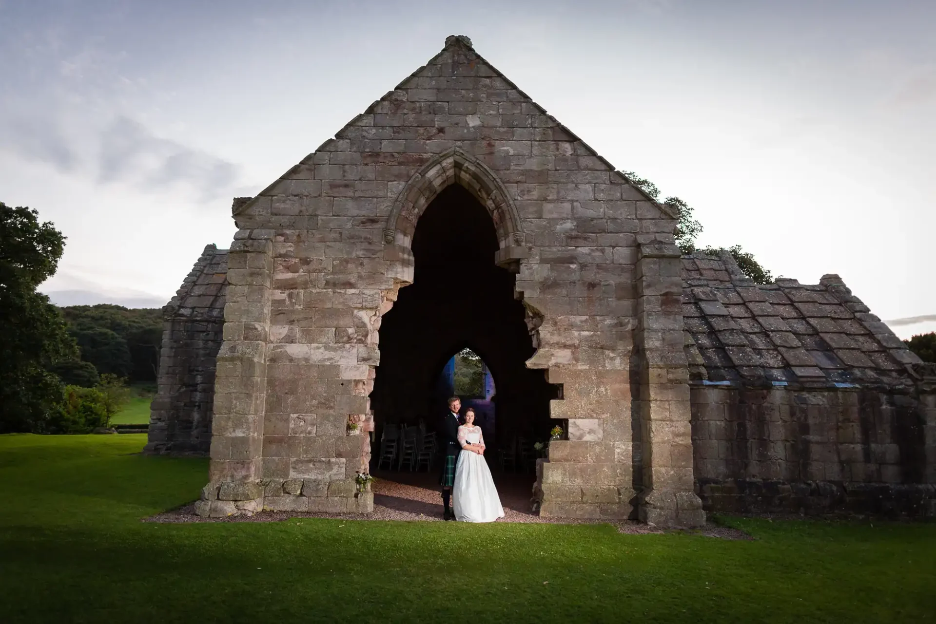 A bride and groom standing in the archway of an old stone chapel with lush greenery in the background at dusk.