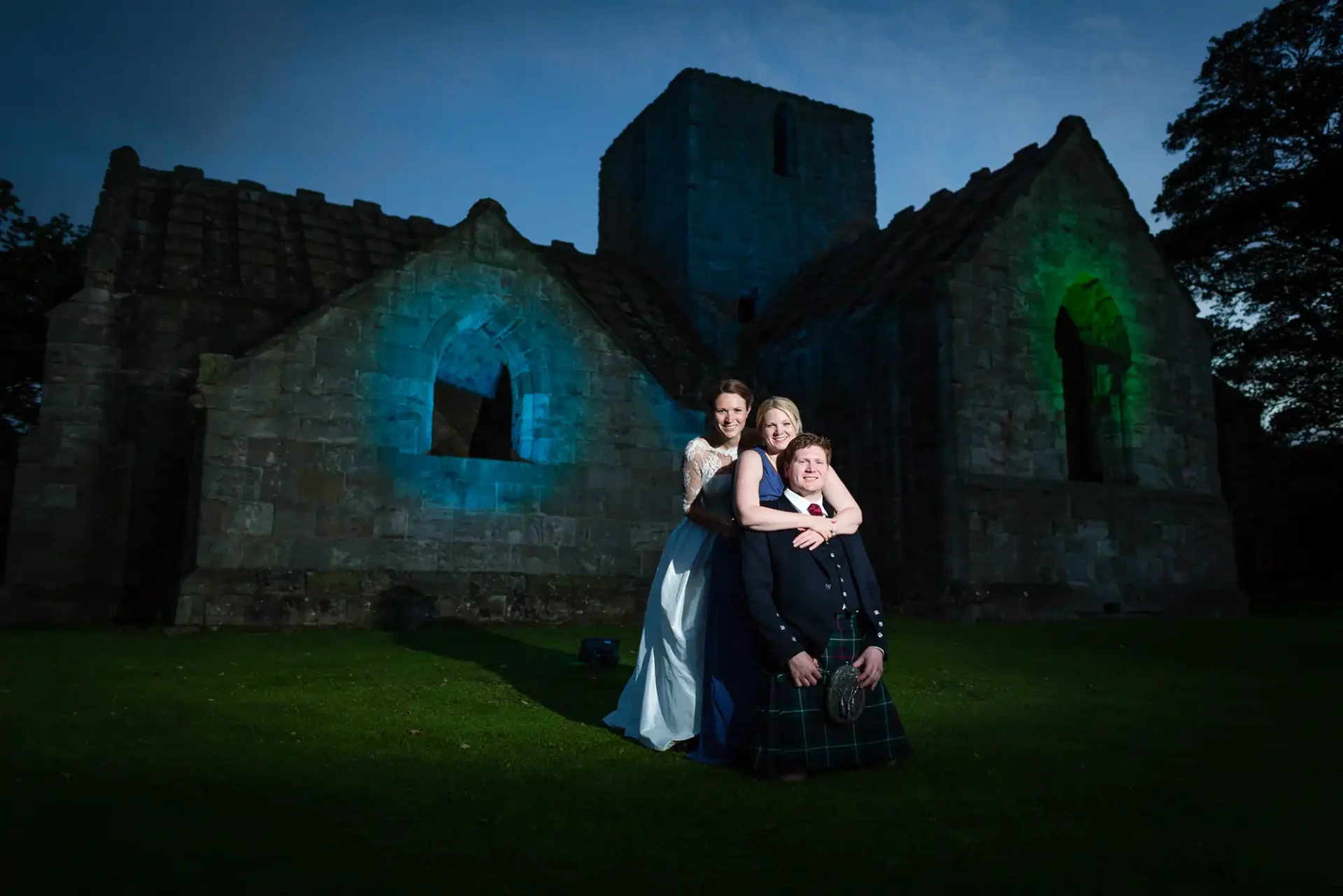 A bride and groom posing at night in front of an old stone church illuminated with blue and green lights.