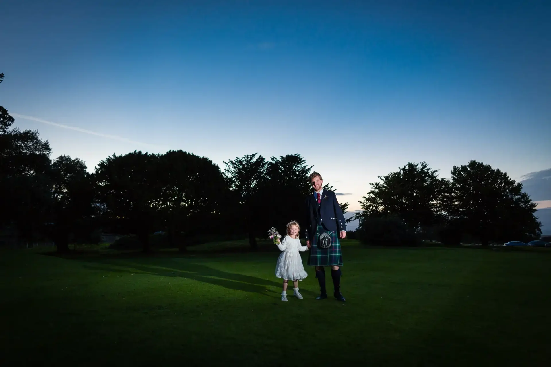 A man in a kilt and a young girl in a white dress standing on a grassy field under a twilight sky.