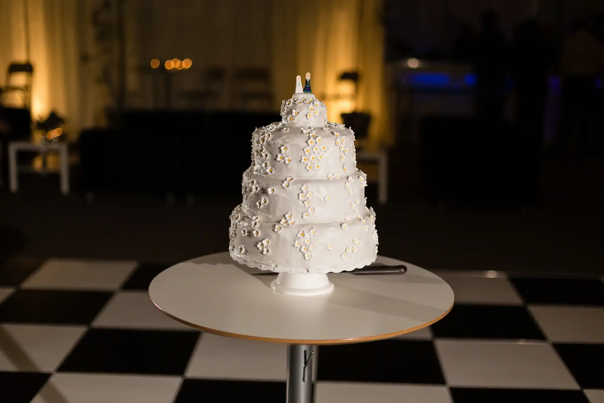 A three-tiered white wedding cake decorated with pearls and gold flowers, displayed on a small table with a checkered floor background.