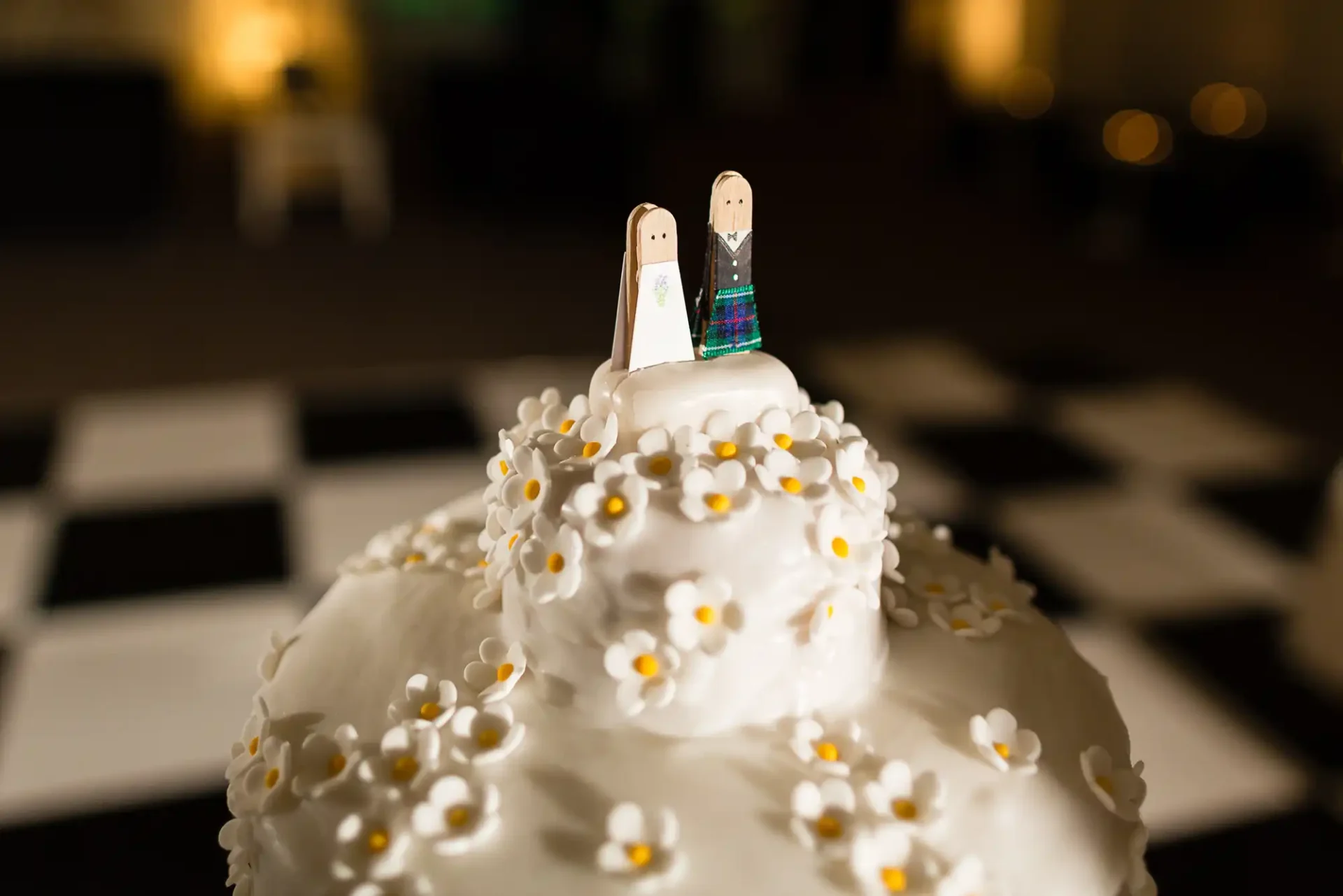 Two wooden figurine toppers on a wedding cake decorated with white flowers, set against a softly blurred background.