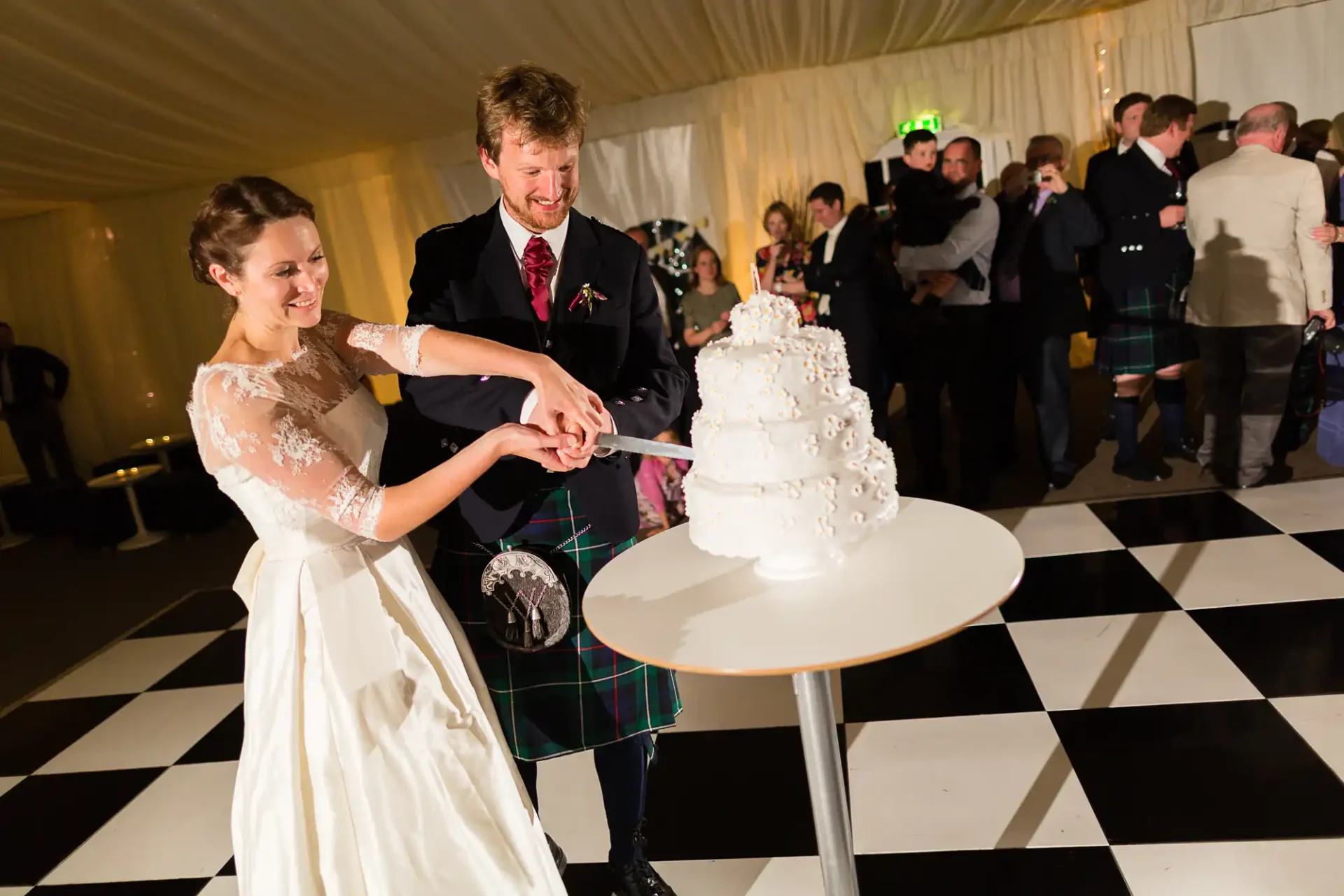 A bride and groom in formal attire, the groom in a kilt, happily cutting a white multi-tier wedding cake at a reception.