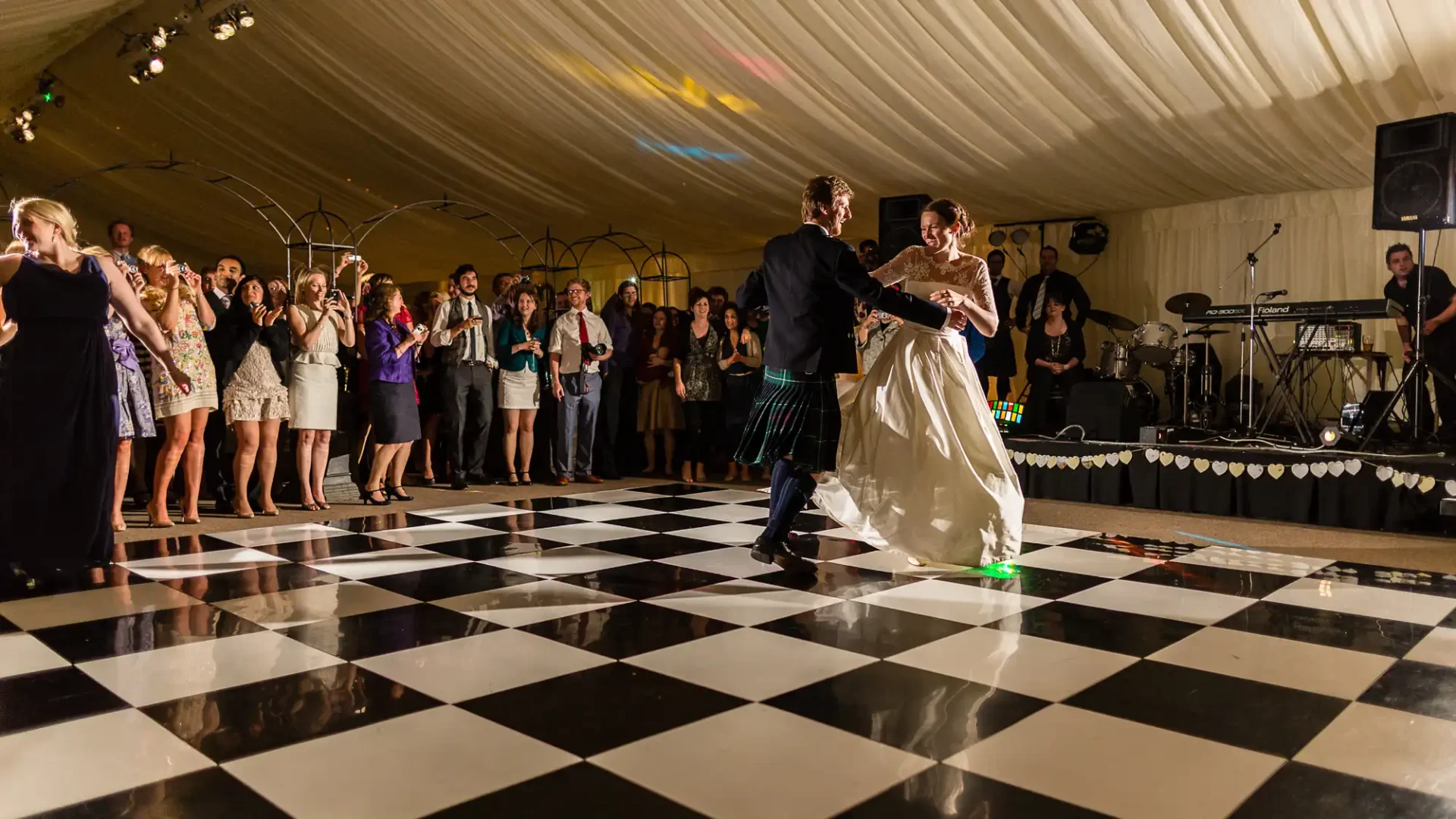 A bride and groom in a kilt dance at their wedding reception, surrounded by cheering guests on a checkered dance floor with a band in the background.