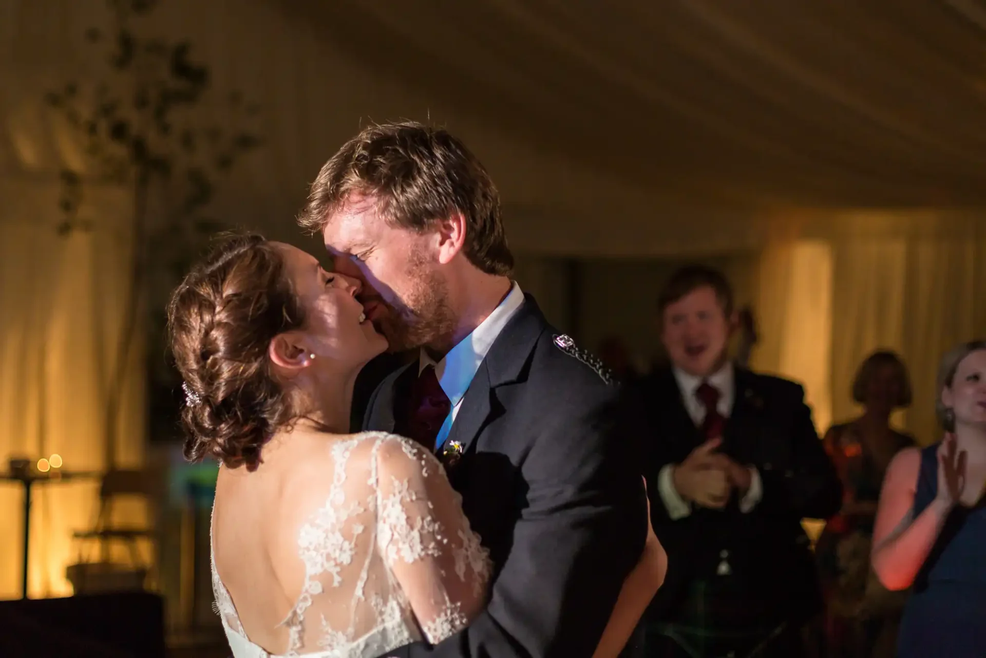 A newlywed couple kissing on the dance floor under a tent, surrounded by applauding guests in soft, warm lighting.