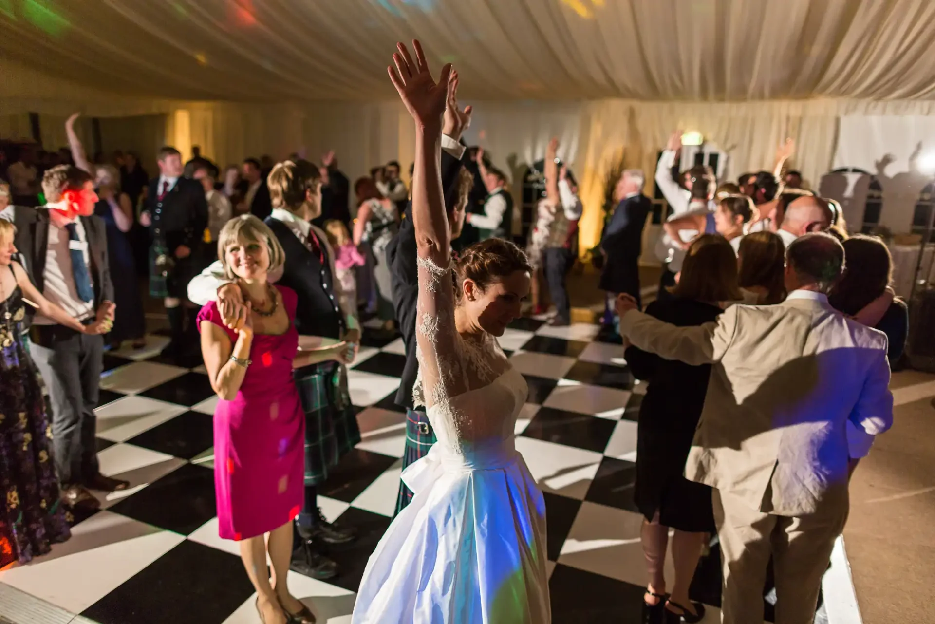 A bride joyfully dancing with raised arms in a crowded reception tent with guests surrounding her, illuminated by warm lights.