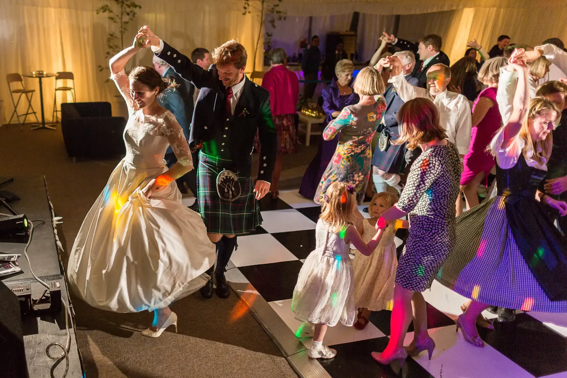 A bride and groom happily dancing with guests in a lively wedding reception hall, illuminated by warm lighting.