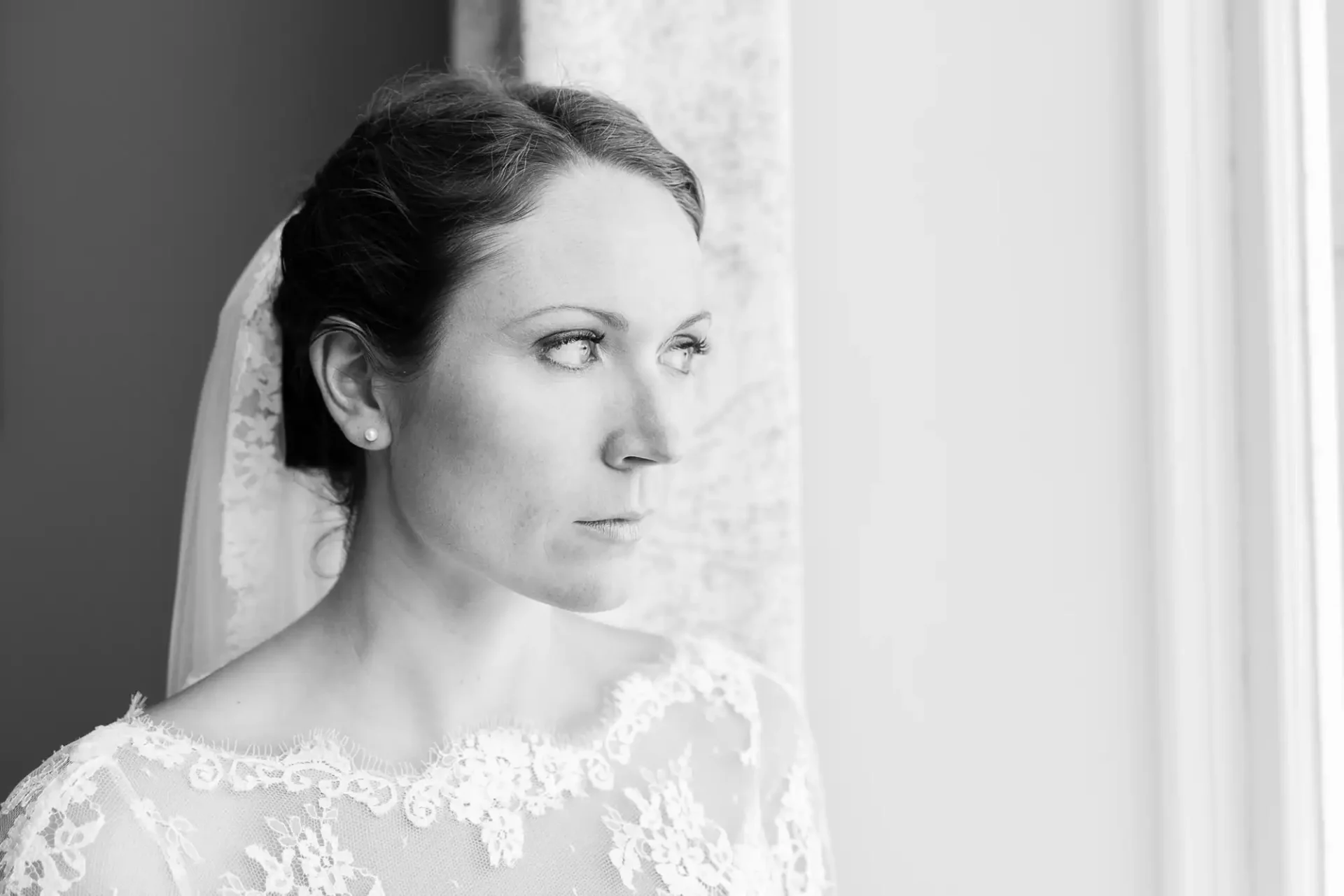 A bride in a lace dress looking out a window in a thoughtful pose, captured in black and white.