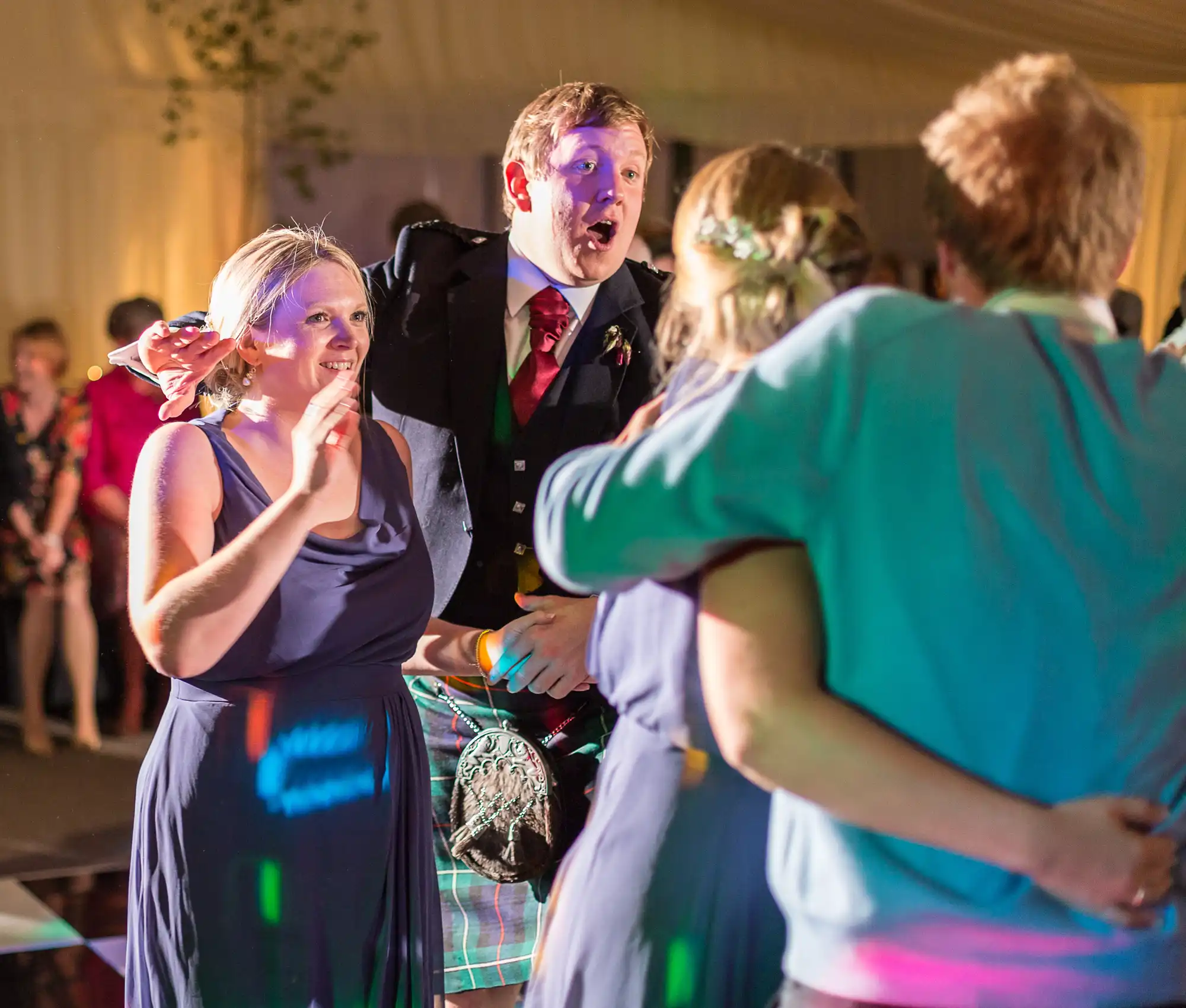 A woman holding a wine glass and a man in a kilt and suit at a tented party engage in a lively conversation with another woman.