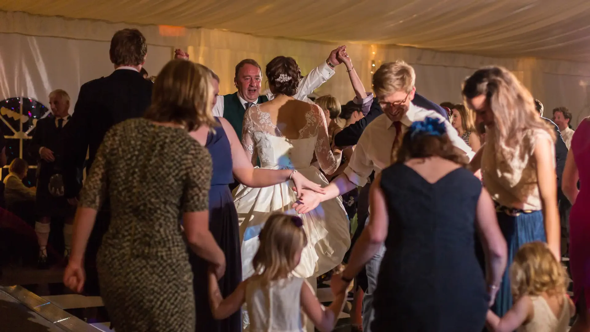 Guests dancing joyously at a wedding reception inside a tent, with a bride in a lacy dress dancing at the center.