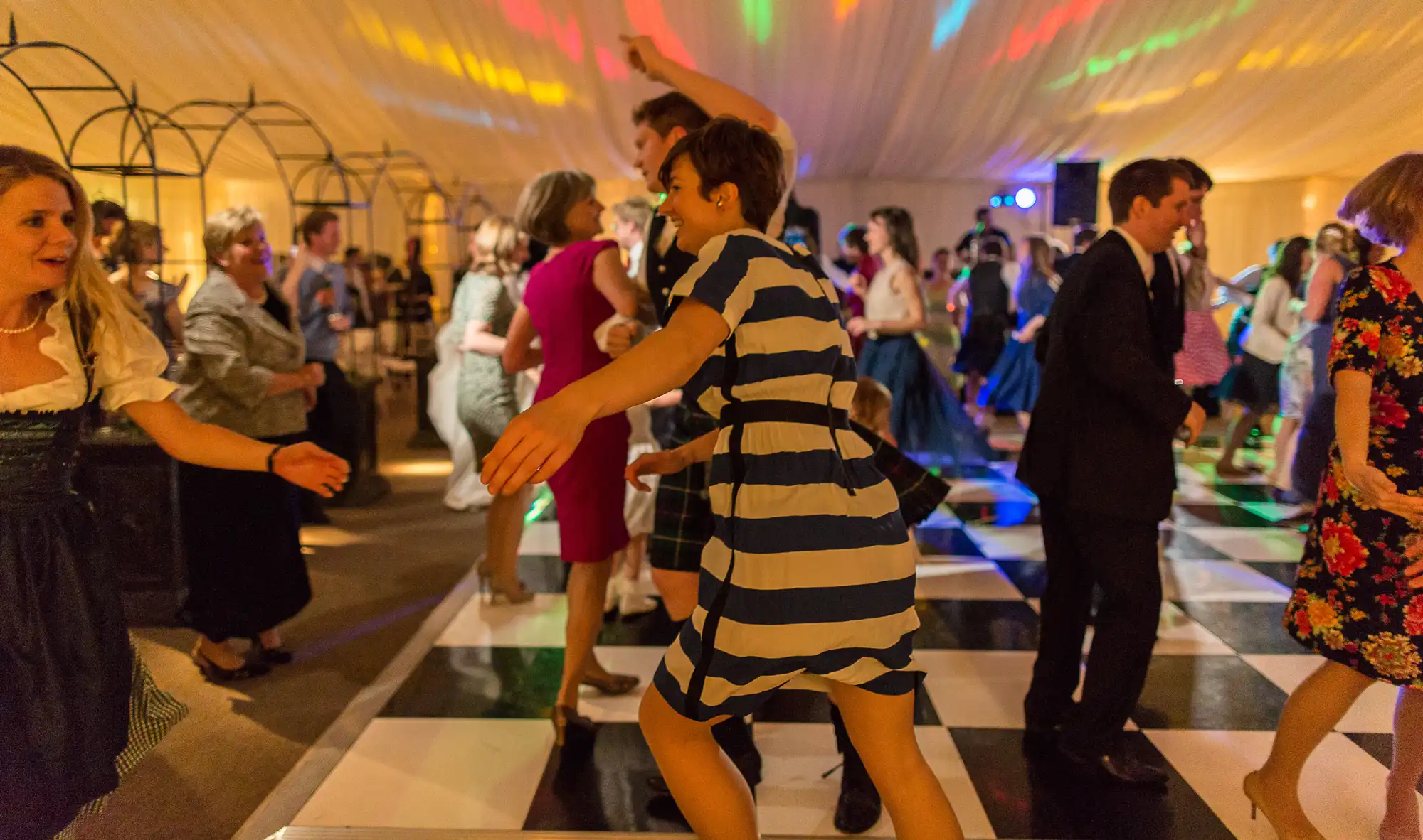 People dancing energetically at a lively party with colorful lights in a tent, some wearing casual and traditional outfits.