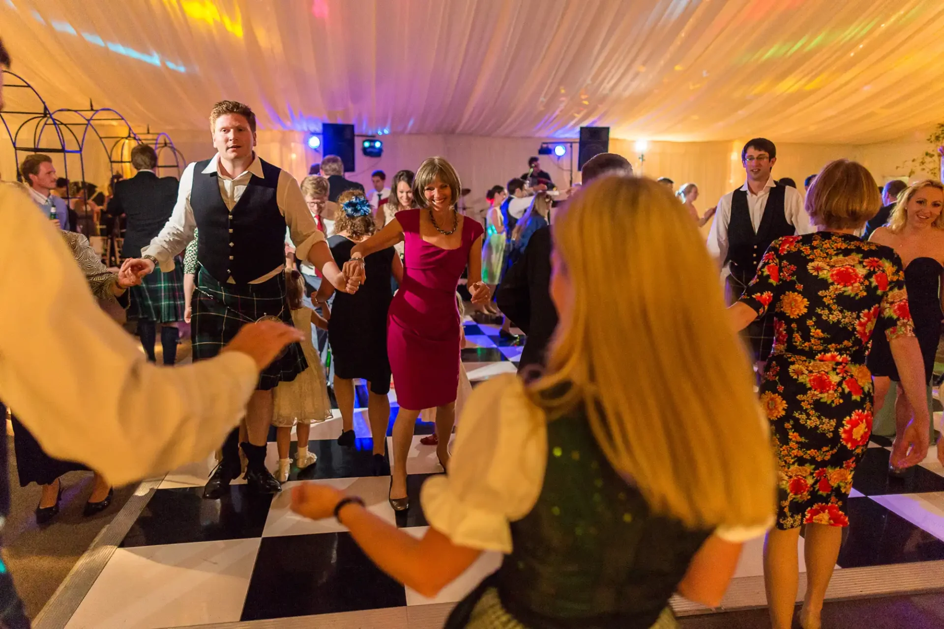 People dancing at a lively indoor party with colorful lights, including a man in a kilt leading the dance.