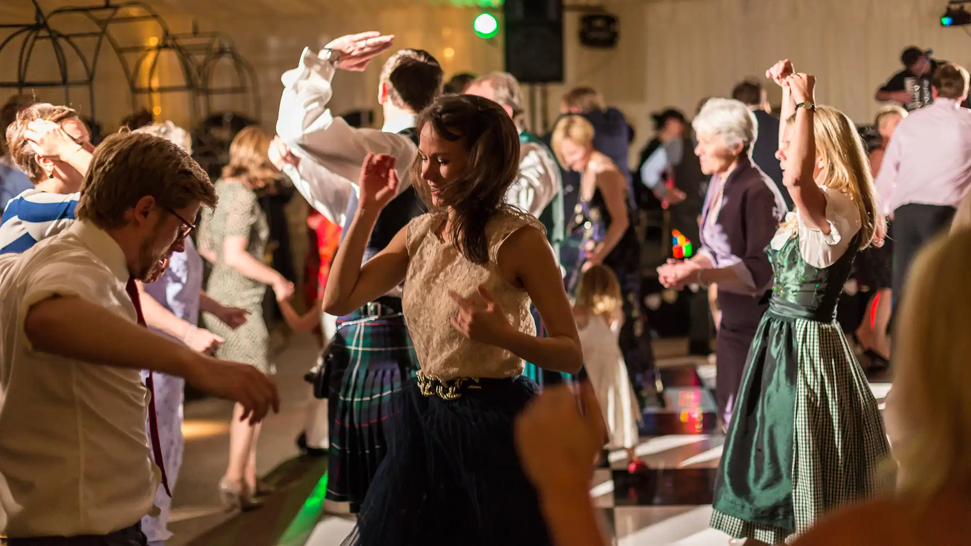 People dancing at a lively event, some wearing traditional costumes, in a warmly lit hall with a wooden floor.