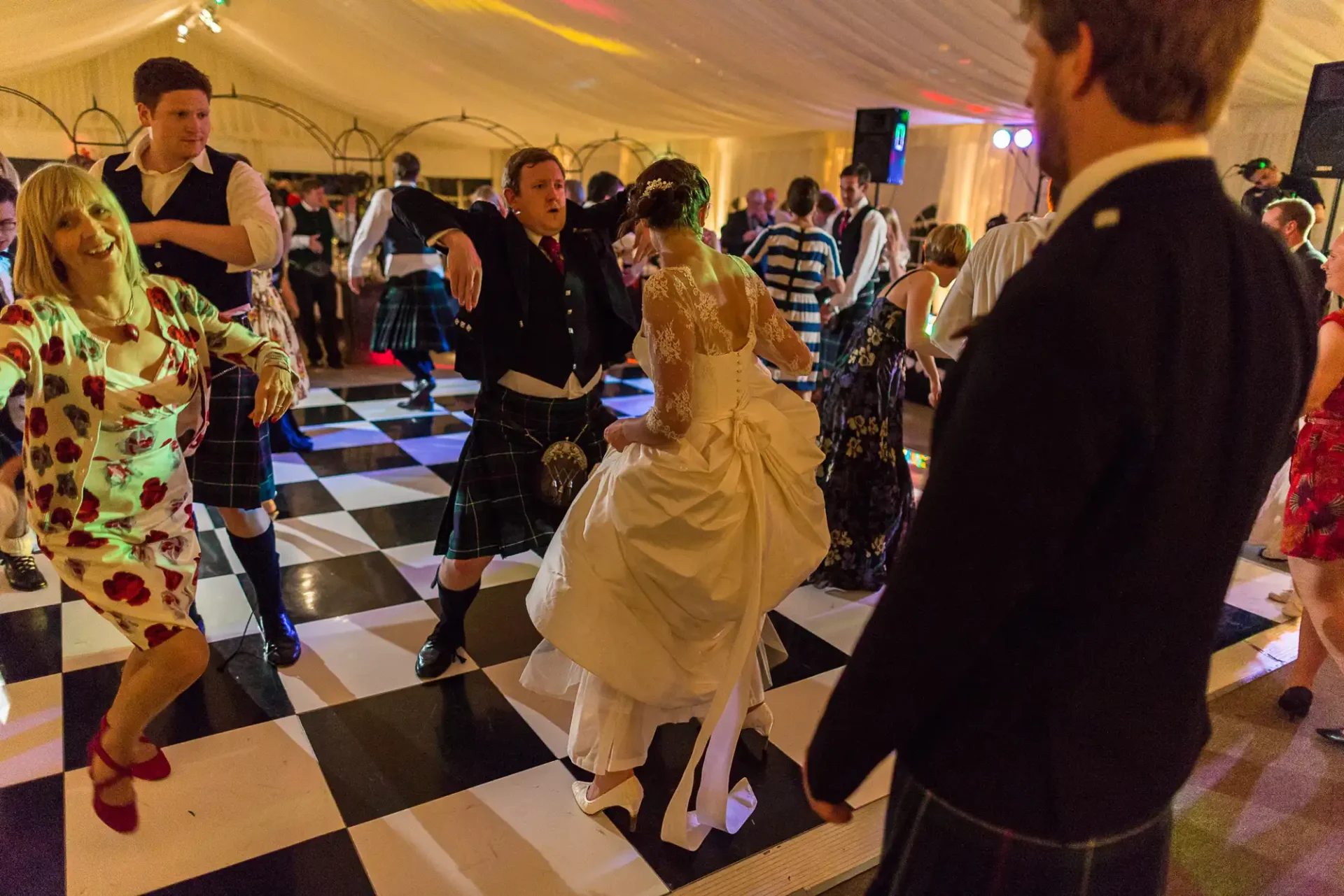 Guests enthusiastically dancing at a wedding reception inside a tent with a checkered floor; a bride in a white dress is in the foreground.