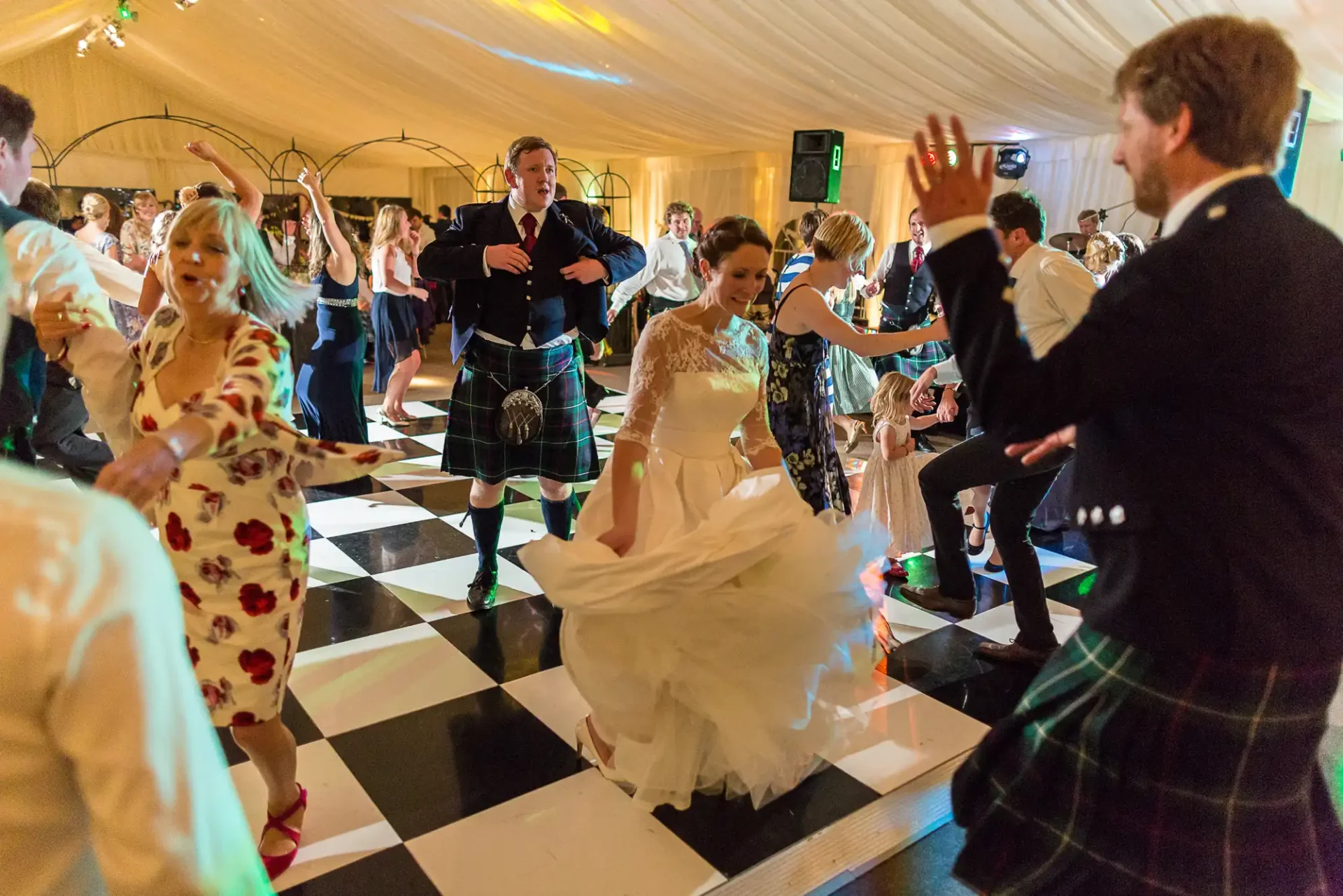 A bride in a white dress dances energetically with guests in kilts and formal attire on a black and white dance floor at a lively wedding reception.