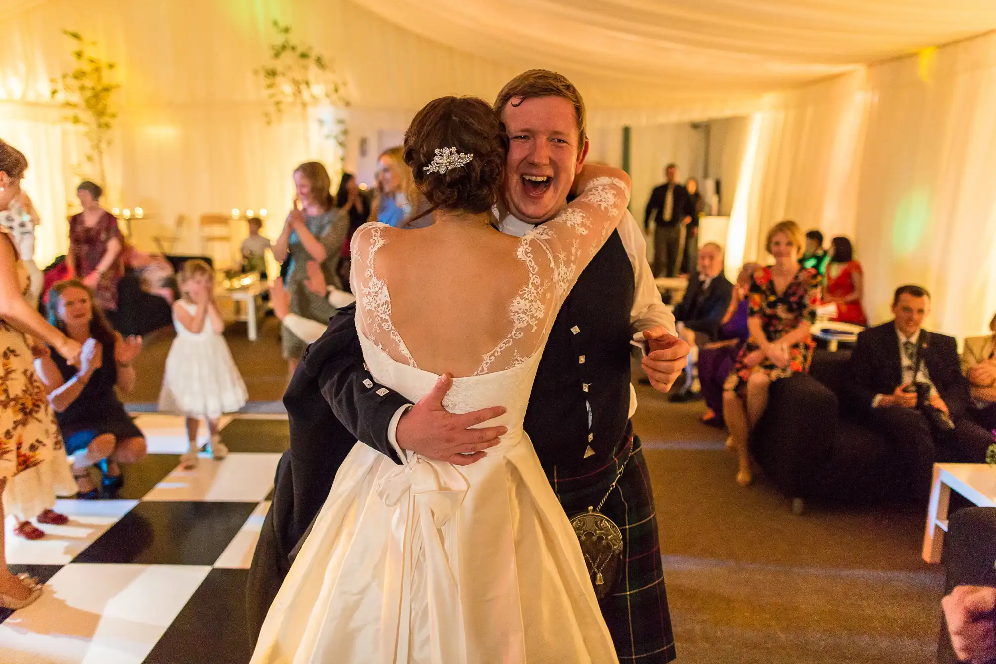 Bride in white gown and groom in kilt hug joyfully on a dance floor at a wedding reception, with guests around them.