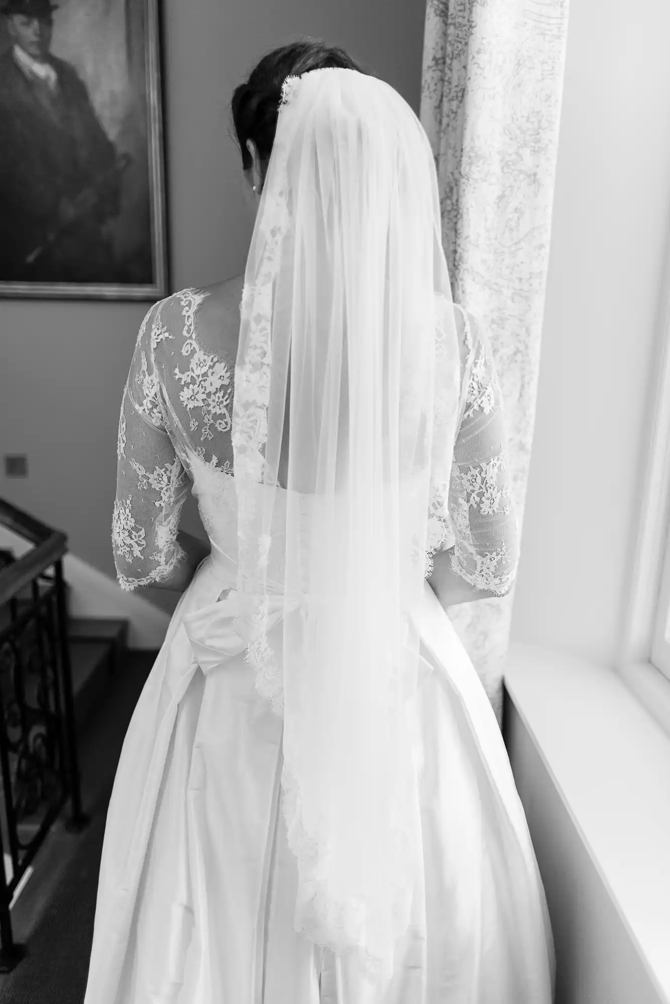 A bride in a lace wedding dress and veil, viewed from behind, looking out a window in a softly lit room.