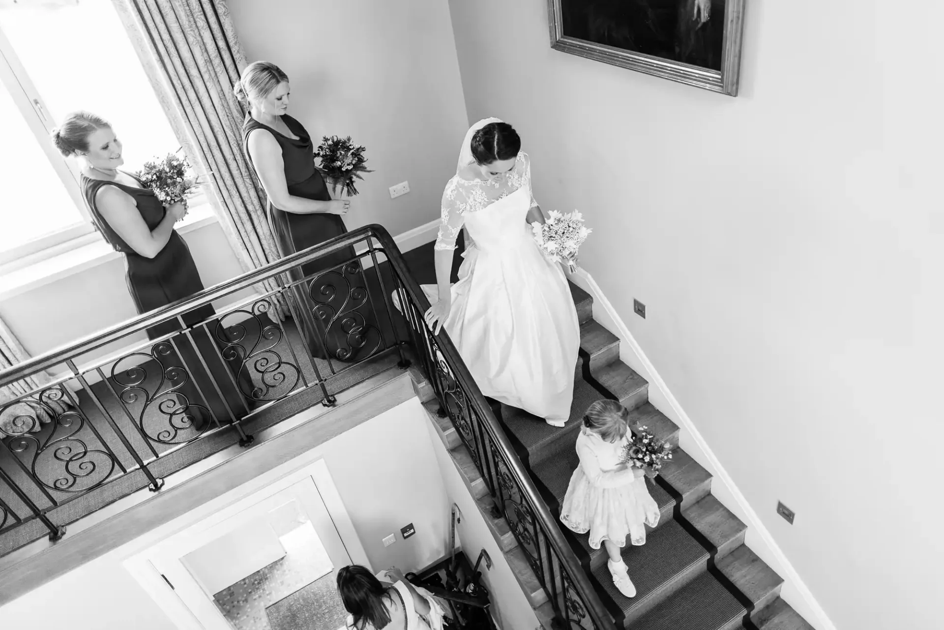 A bride in a white gown descends a staircase followed by bridesmaids and a young flower girl in a bright room, captured in black and white.