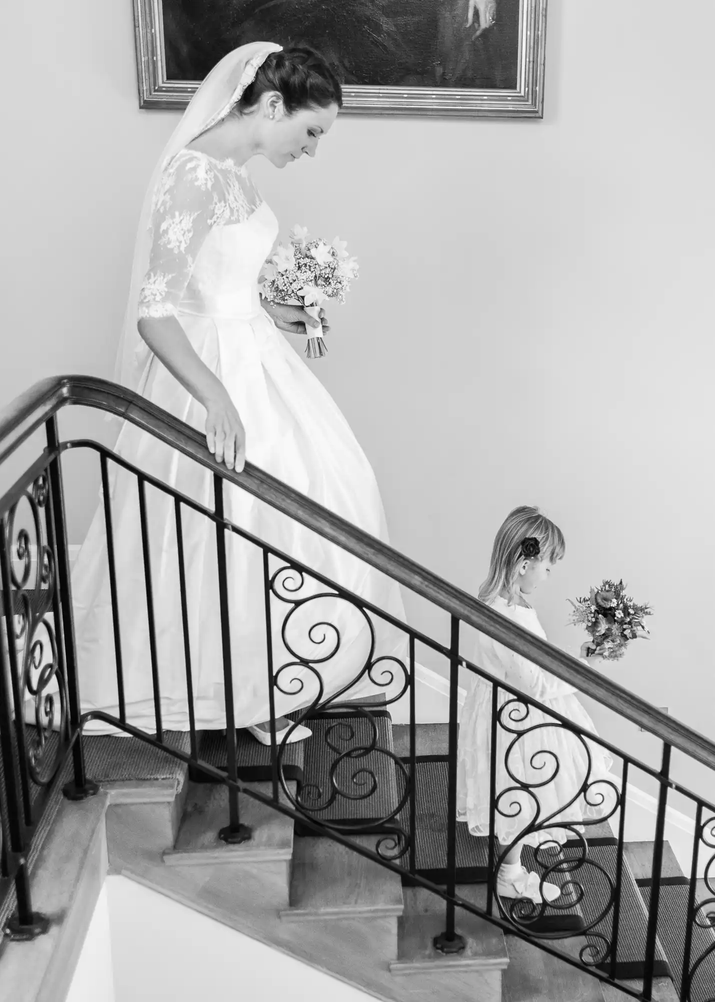 A bride in a white dress descends a staircase, smiling at a young girl holding a bouquet at the bottom. black and white photo highlighting elegant details.