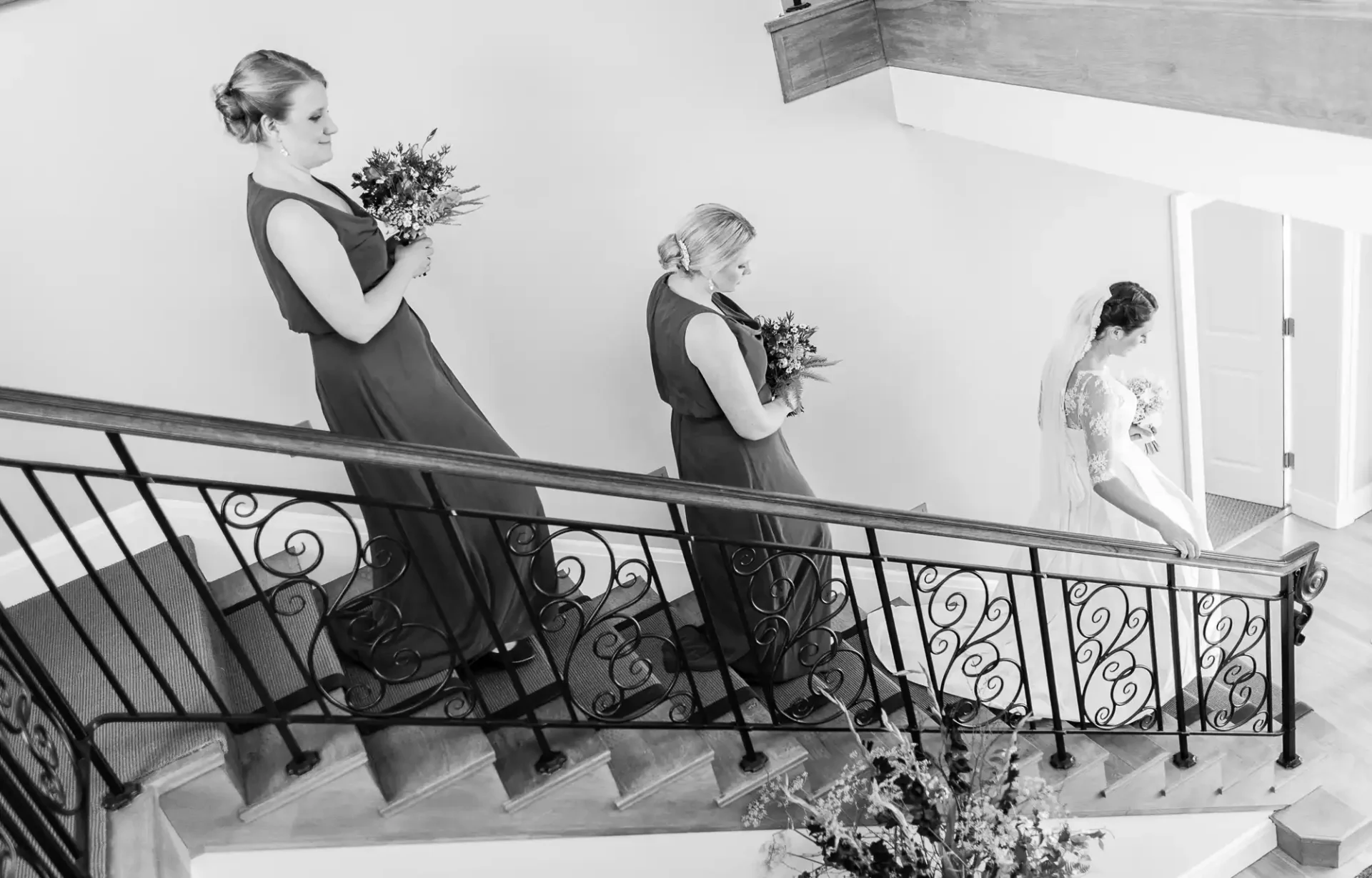 Three bridesmaids in dark dresses carry bouquets, descending a staircase with ornate railings, with a bride in white visible in the background.