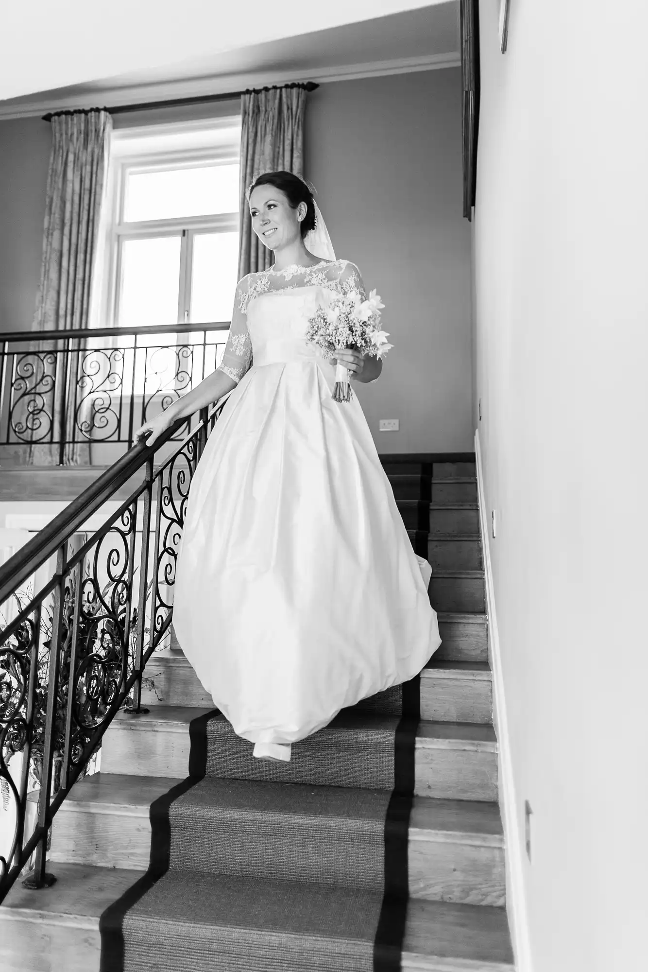 A bride in a traditional white dress holding a bouquet smiles as she descends a staircase in a well-lit room.