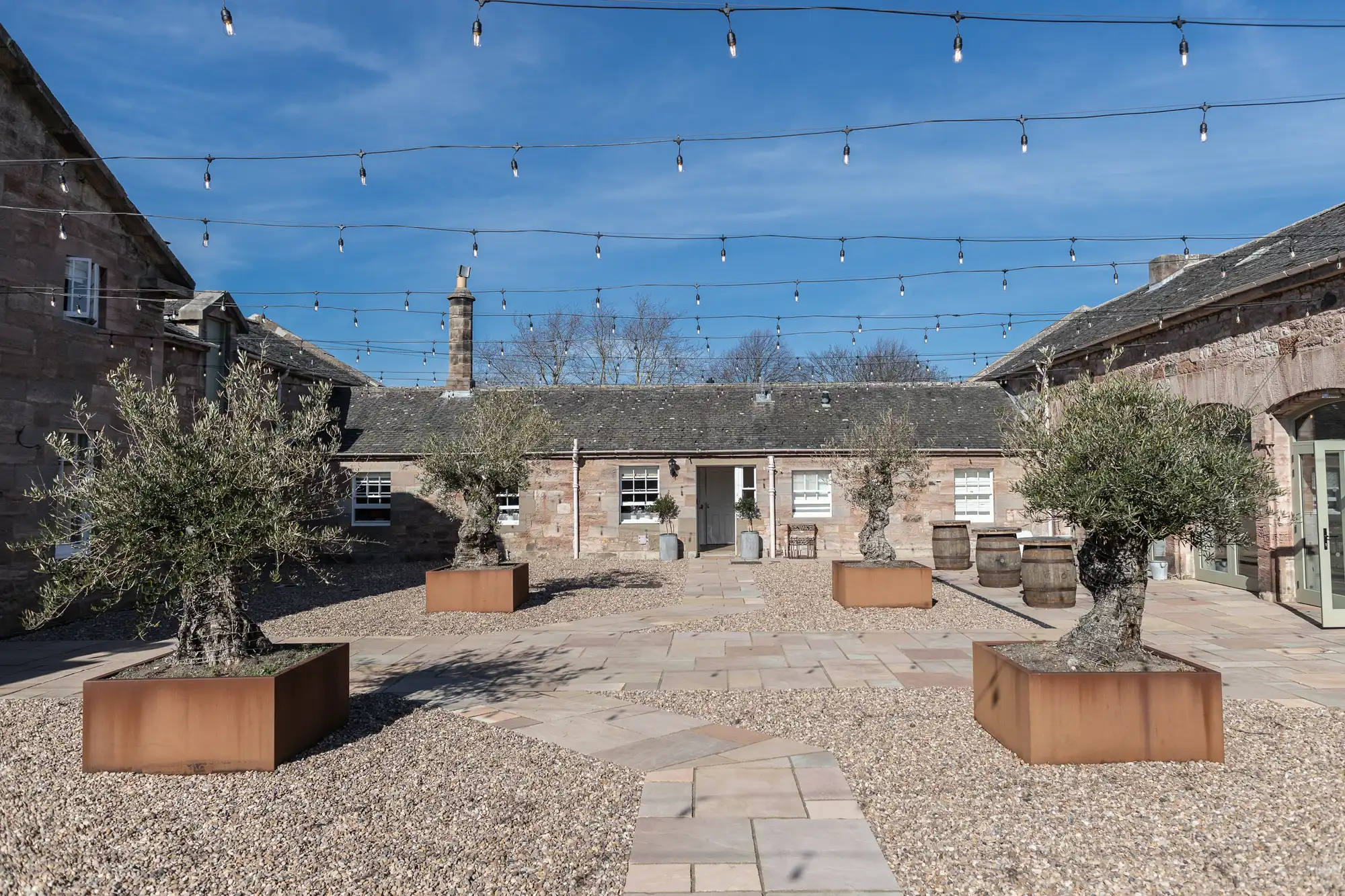 Courtyard with stone buildings, potted trees, and string lights overhead, set under a clear blue sky.