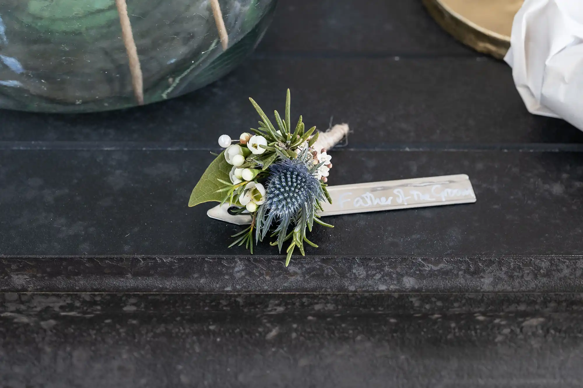 A boutonniere featuring a blue thistle, white berries, and green foliage lies on a dark surface alongside a name tag reading 'Father of the Groom.'.