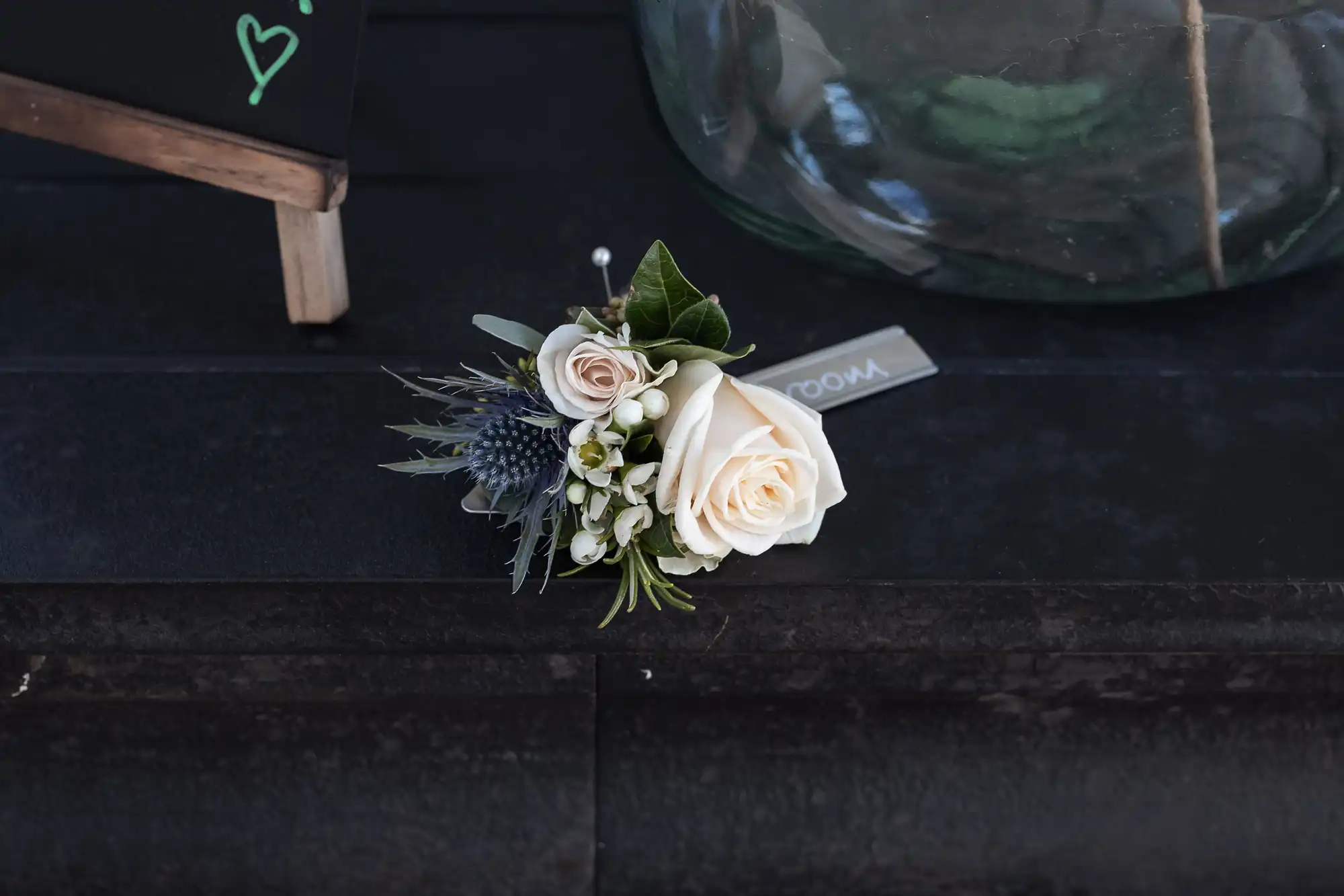 A small boutonniere with a white rose, white buds, and greenery is placed on a black surface next to a glass container.