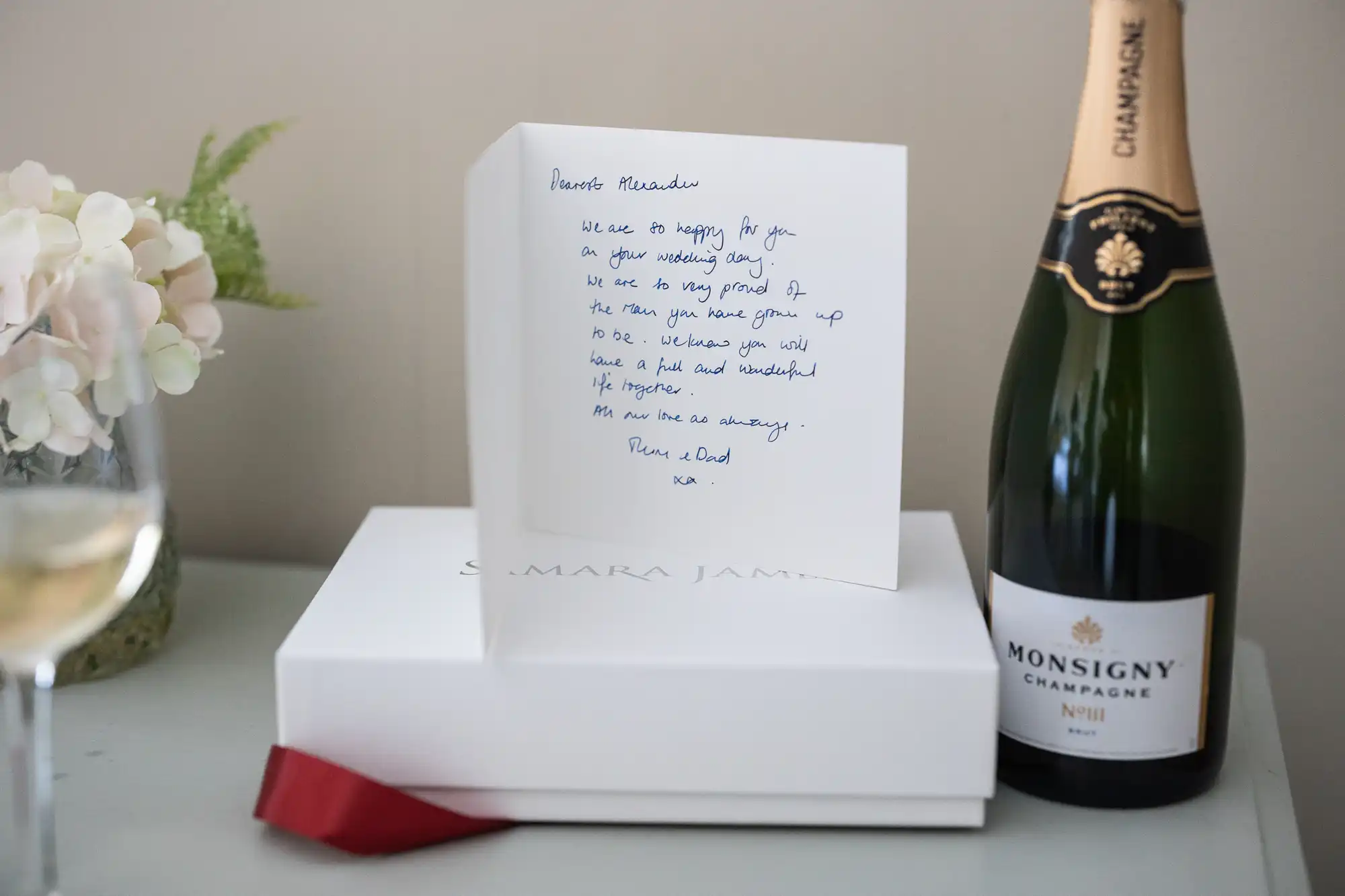 A handwritten card stands open near a bottle of Monsigny champagne and a white box with a red ribbon. A glass of white wine is visible to the left. Flowers are in the background.