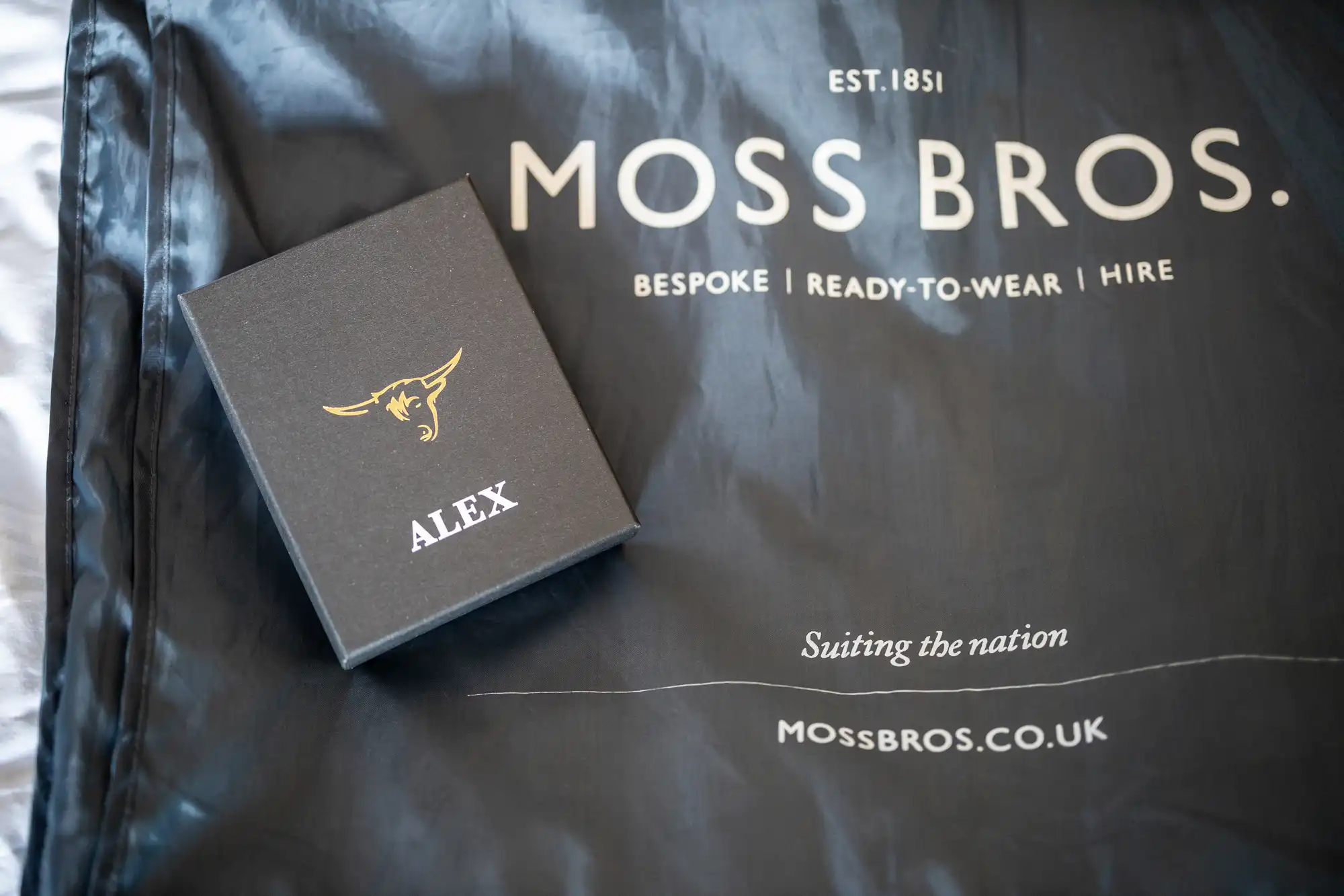 A black box with "ALEX" and a bull emblem on it is placed on a Moss Bros. garment cover. The text on the cover reads: "Moss Bros. Bespoke | Ready-to-Wear | Hire. Est. 1851. Suing the nation. mossbros.co.uk".