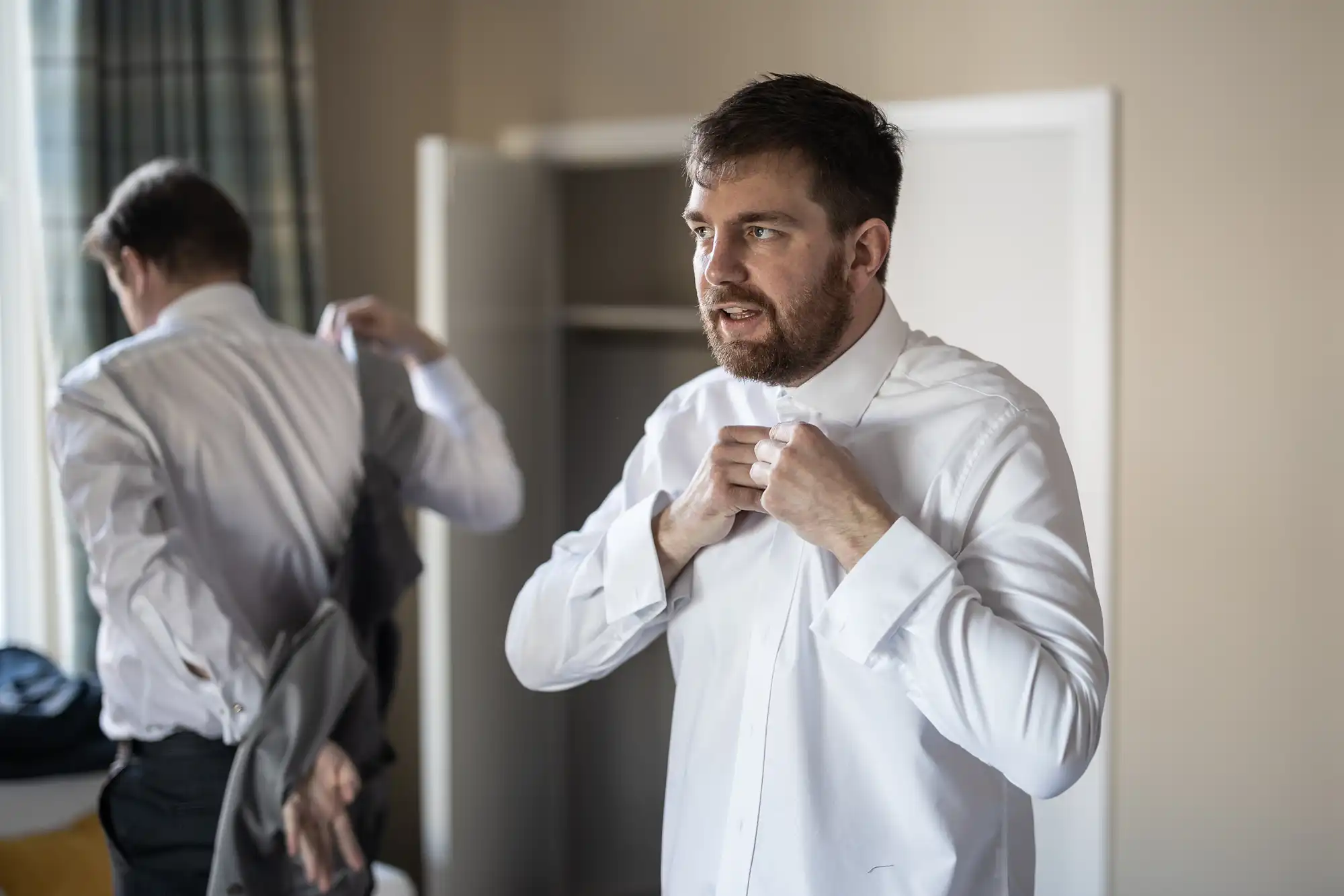 Two men are seen indoors, both wearing white shirts. One man is adjusting his necktie while the other, reflected in a mirror, appears to be putting on a jacket. The background includes a partially open door.