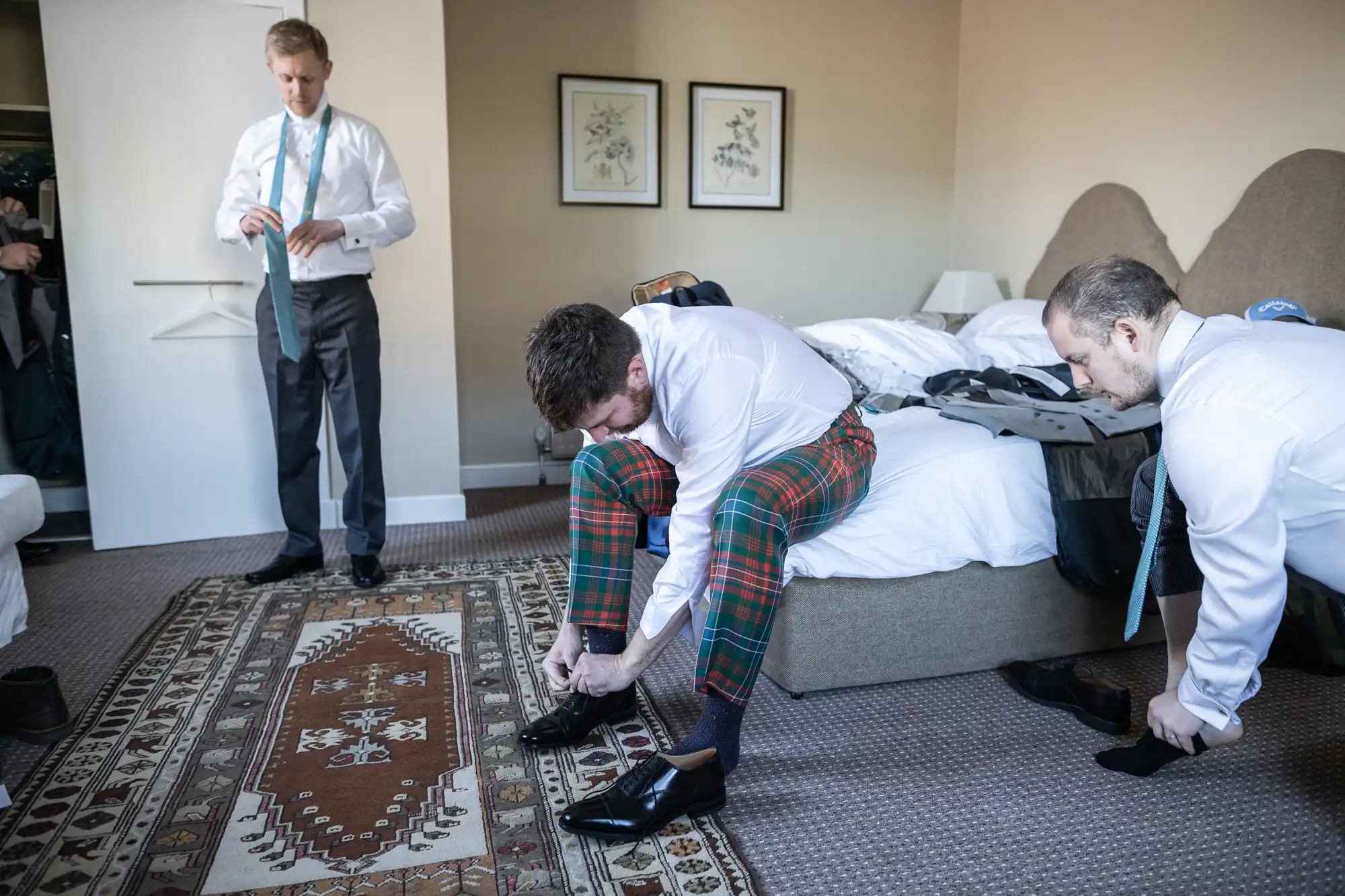 Three men in a bedroom wearing white shirts. One is tying his shoe while wearing plaid pants, another is putting on socks, and the third is adjusting his tie in front of a mirror.