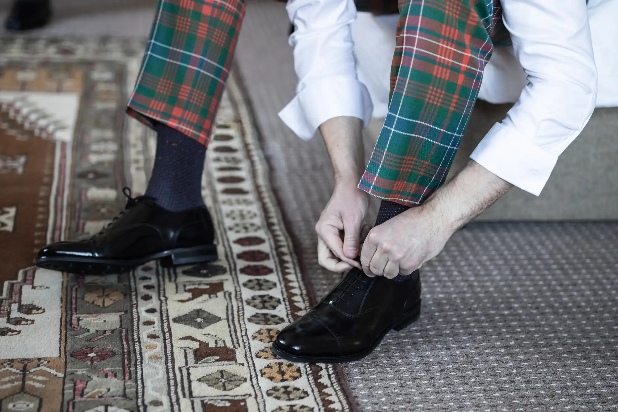 A person wearing plaid pants ties the laces of a black dress shoe while kneeling on a patterned rug.