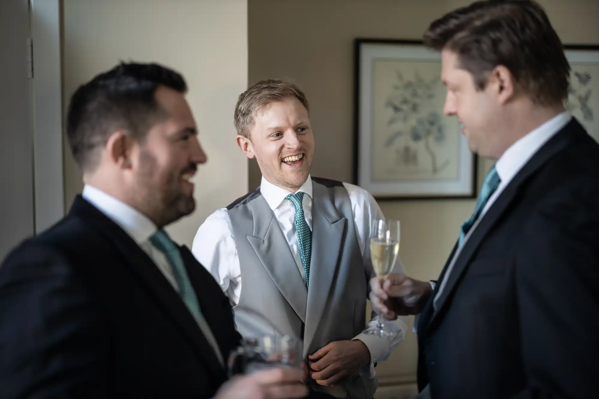 Three men in formal attire, one in a grey vest and white shirt, and two in black suits, are standing and smiling while holding drinks, in a room with framed art on the wall.