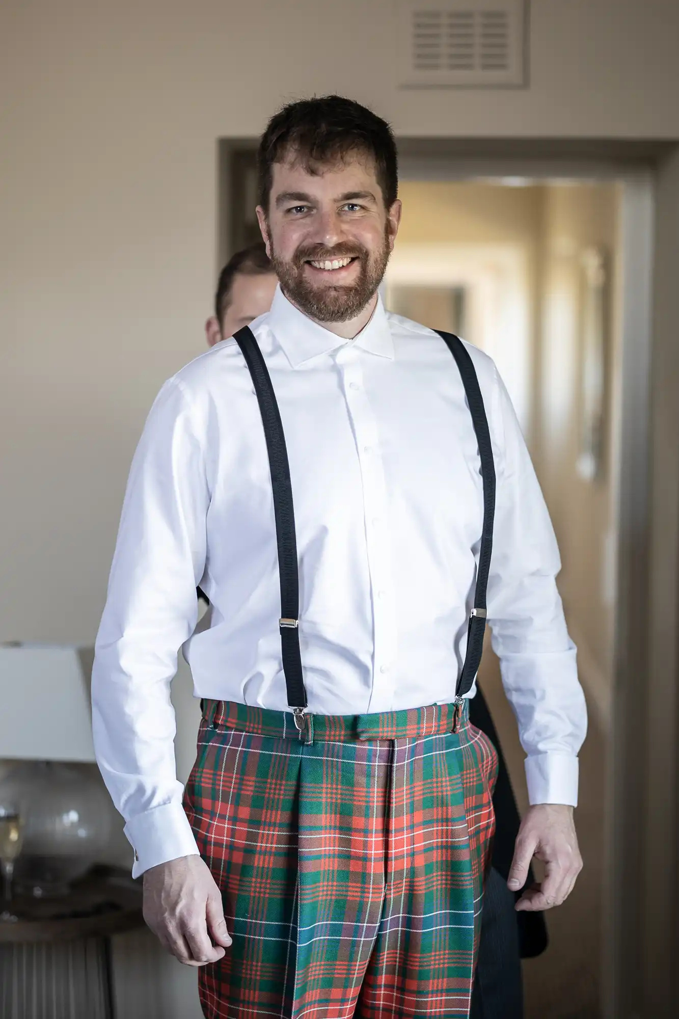 Man wearing a white dress shirt with black suspenders and tartan plaid pants, smiling indoors.