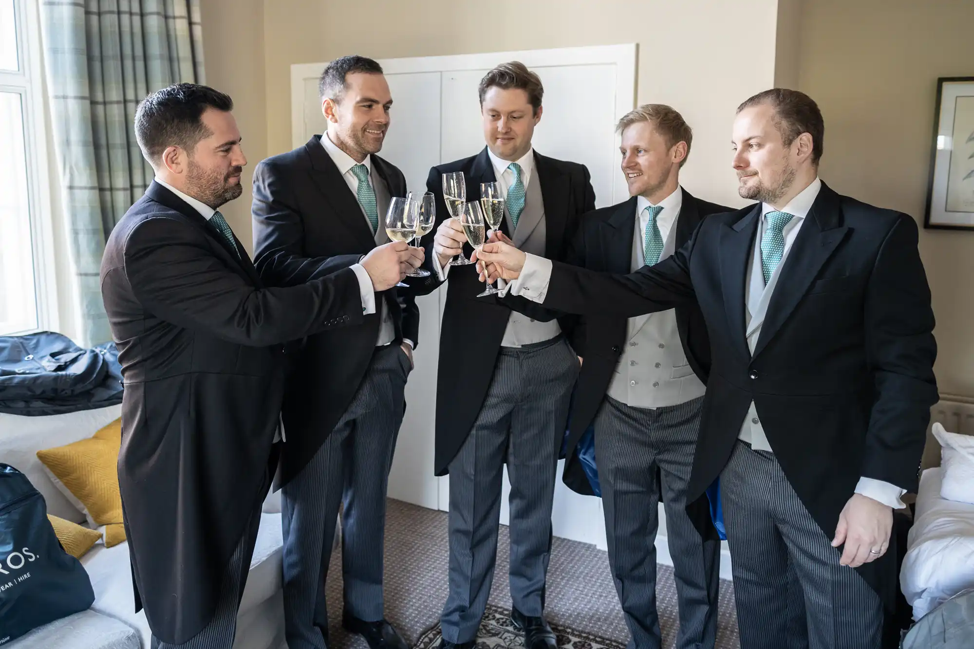 Five men in formal wear, holding glasses, making a toast in a well-lit room with a window and a white door in the background.