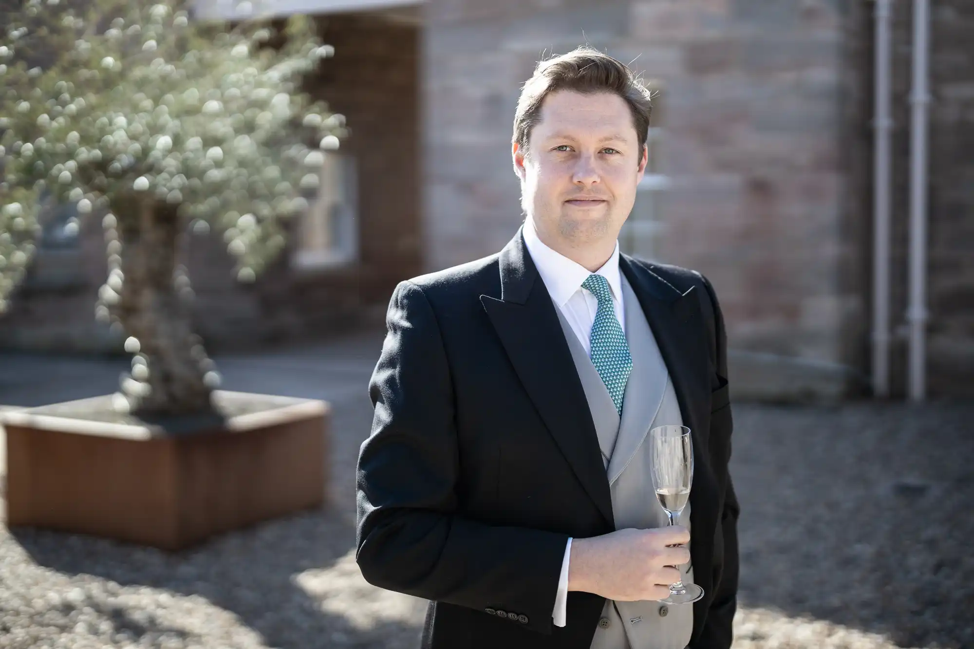 A man dressed in formal attire holds a champagne glass, standing outdoors in a courtyard with a potted plant and stone building in the background.