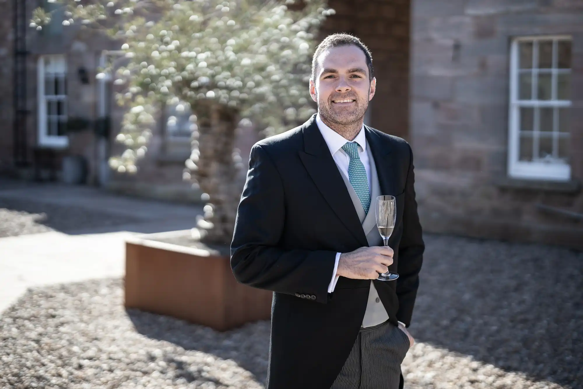 A man in a suit and tie holds a glass of champagne outdoors beside a brick building with a large potted plant in the background.