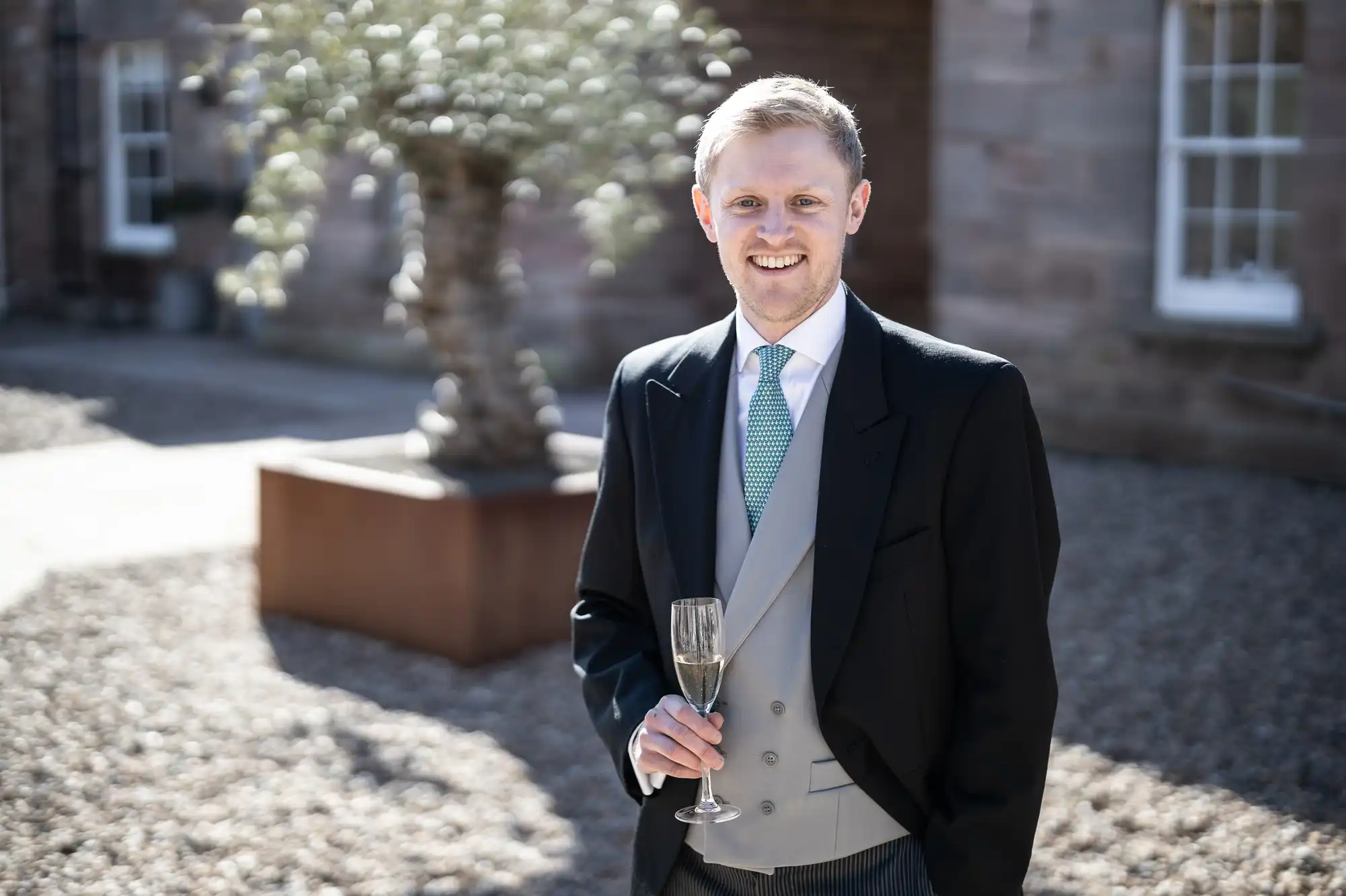 A man in formal attire smiles while holding a champagne glass. He stands outdoors with a blurred background featuring a potted tree and a building.