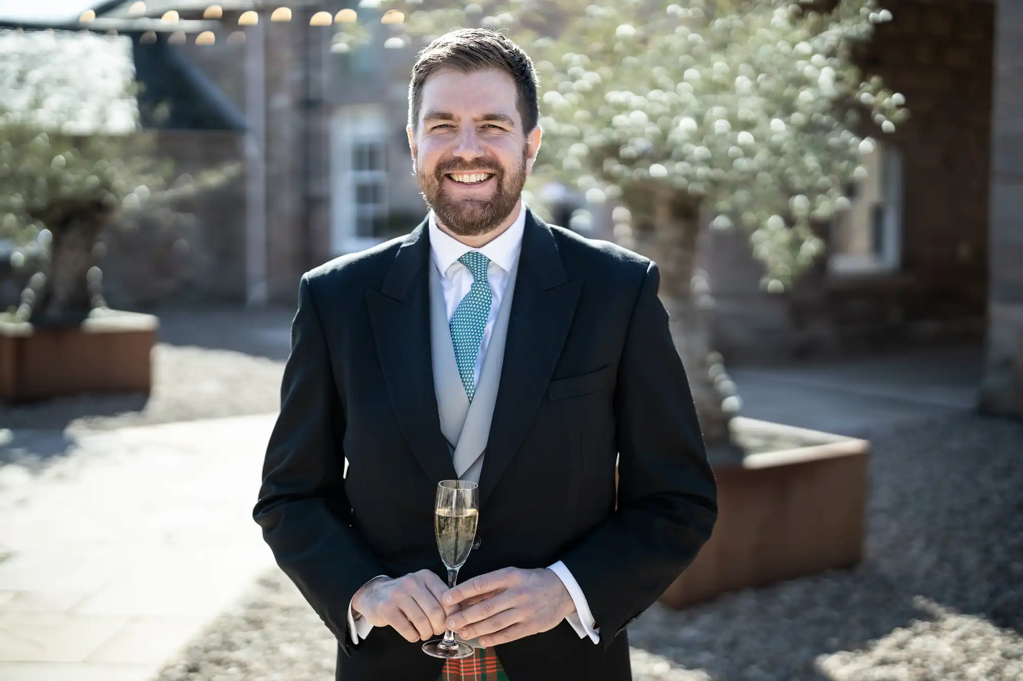 A man in a suit with a blue tie holds a glass of champagne. He is standing outside near buildings and trees.