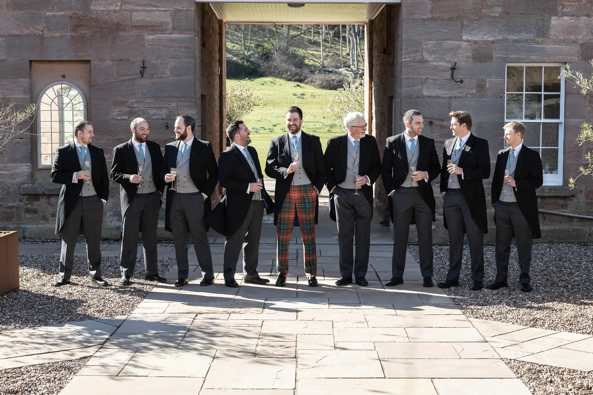 A group of nine men, dressed in formal attire, stands in a line holding drinks. One man in the center wears red tartan pants. They are outside a stone building with an arched doorway.