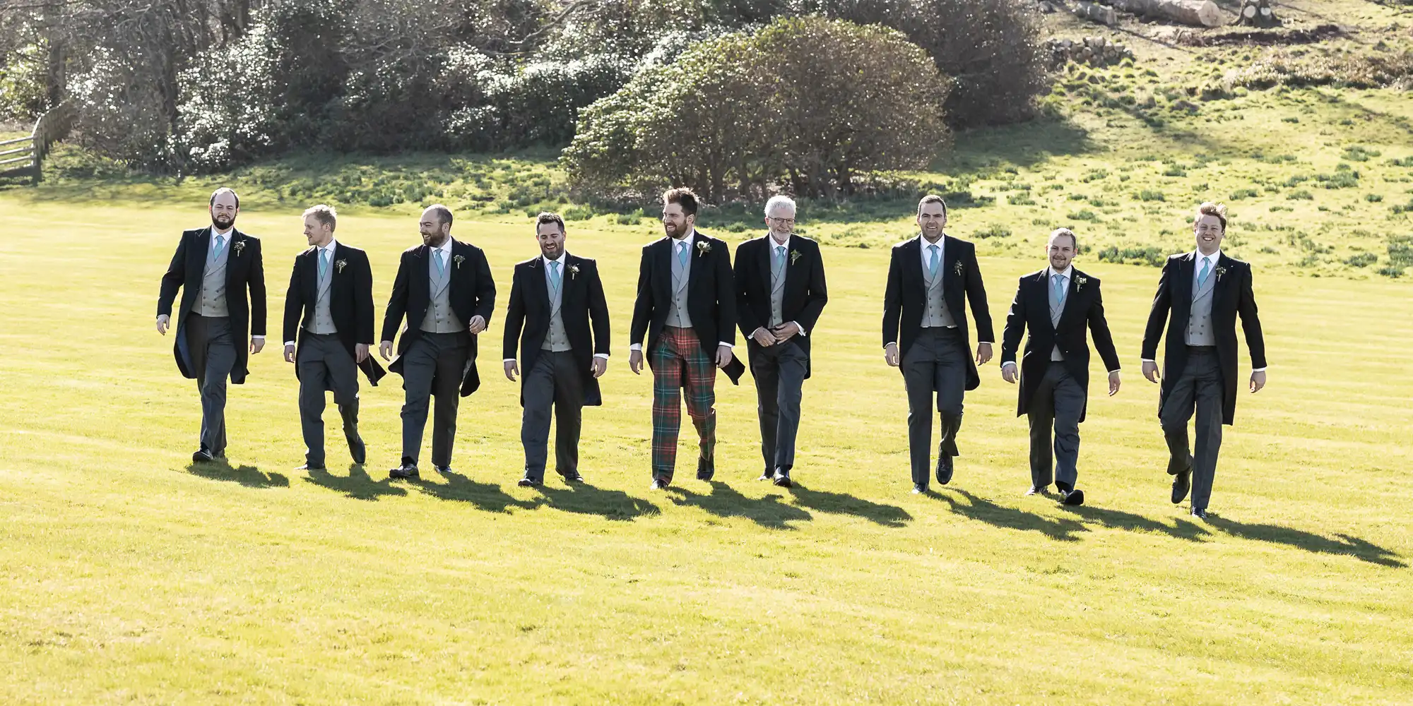 Eight men dressed in formal attire, one in plaid pants, walking across a grassy field with trees in the background.