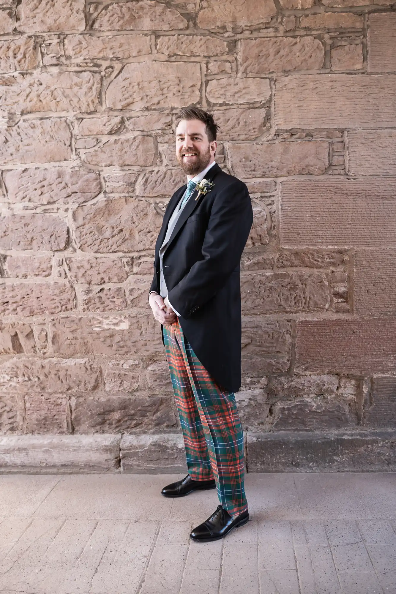 A man stands in front of a stone wall, wearing a black jacket, green tie, and plaid tartan trousers, smiling with his hands clasped.