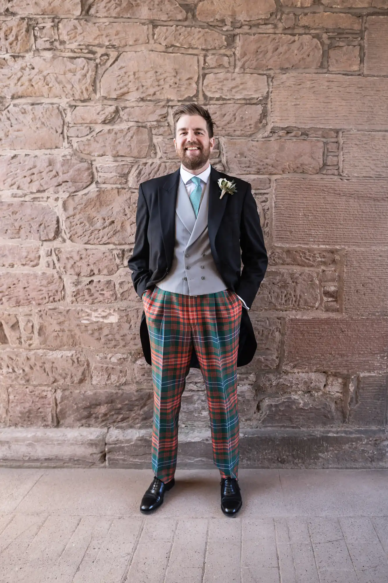 Man in a formal outfit with a black coat, grey vest, white shirt, teal tie, tartan pants, and black shoes stands against a stone wall. He is smiling with his hands in his pockets.