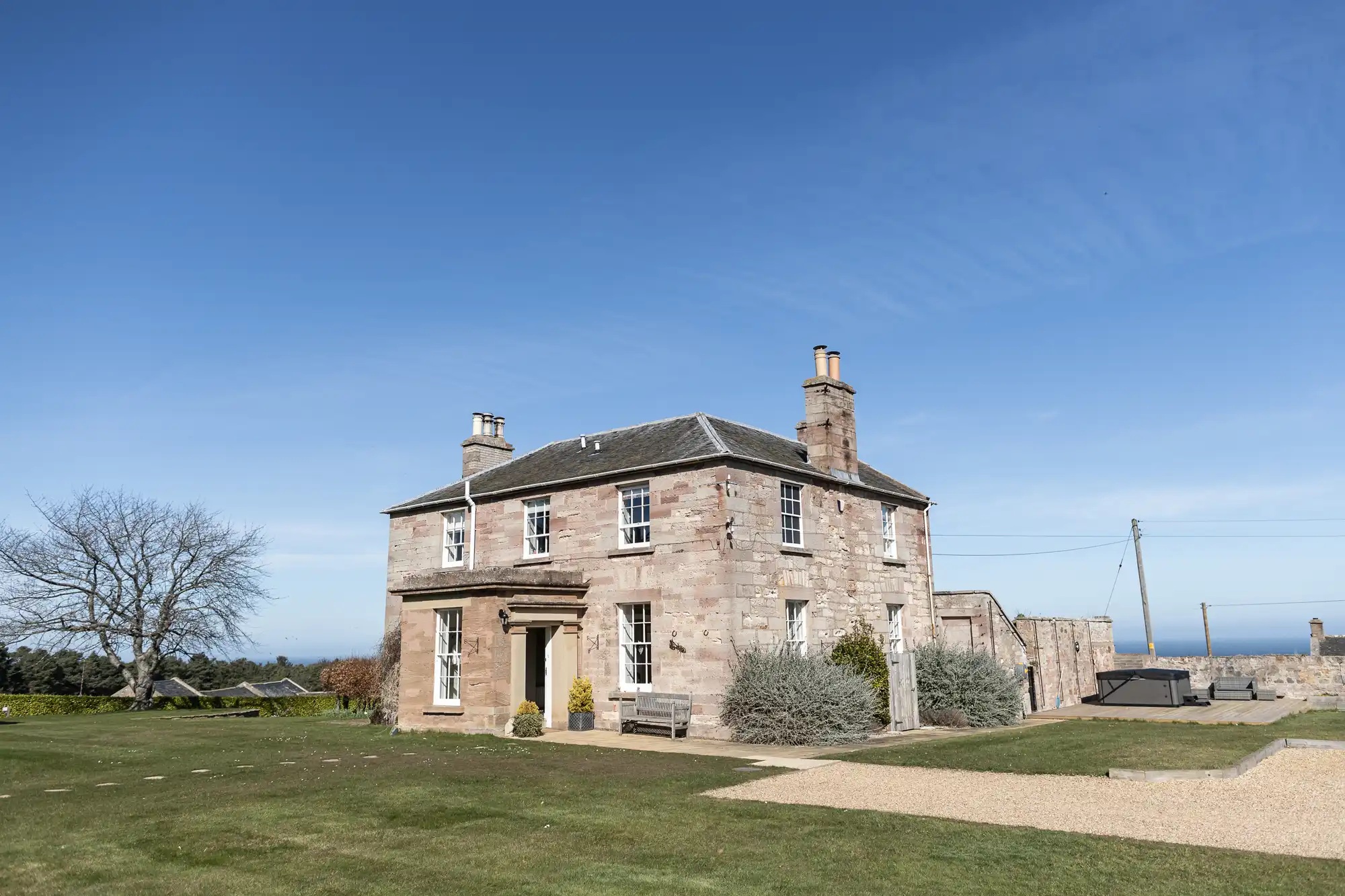 A two-story stone house with chimneys on a clear day, surrounded by a grassy lawn, a tree on the left, and a gravel path on the right.