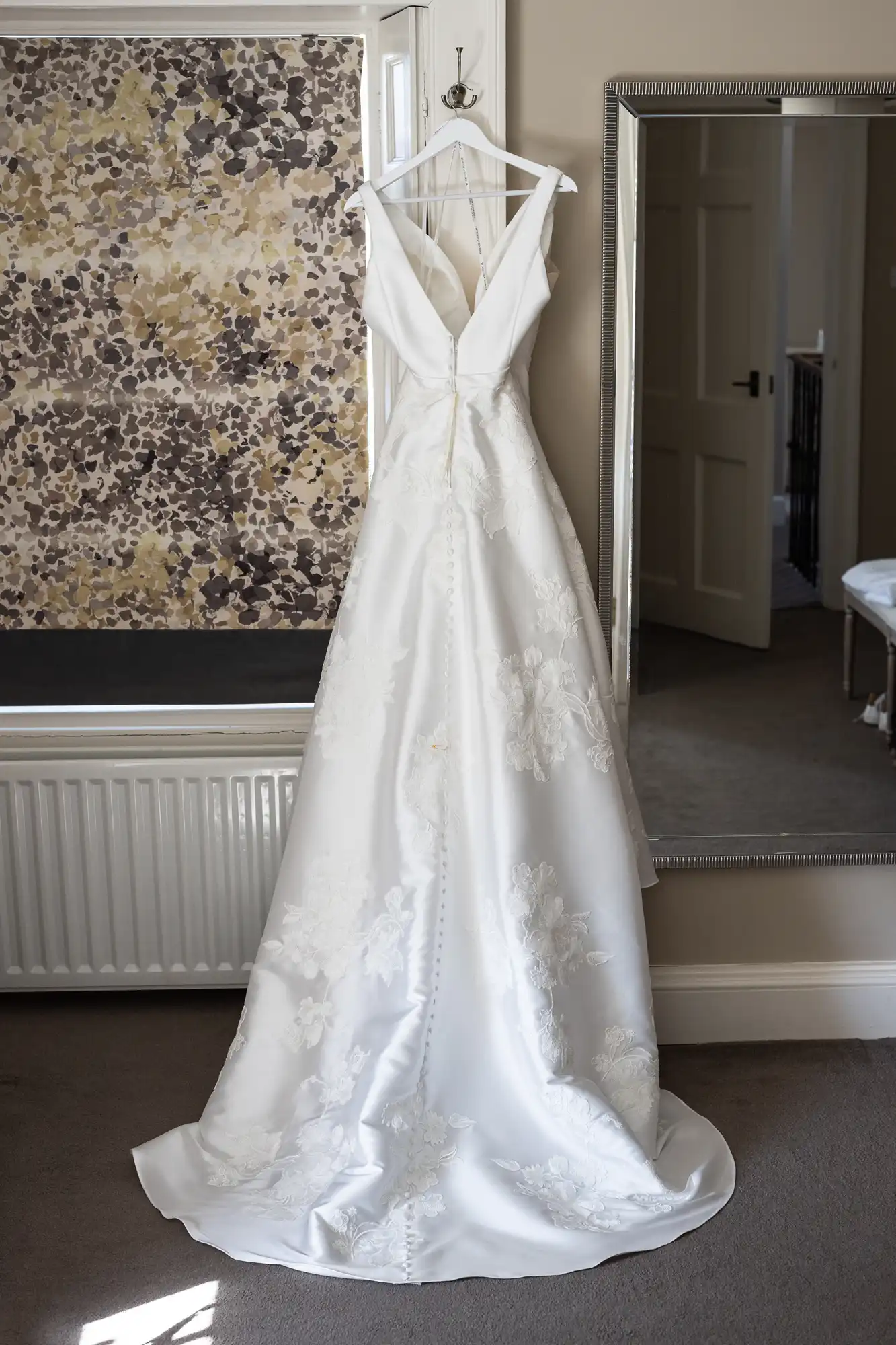 A white wedding dress with a deep V-neck and floral lace detailing hangs on a hook against a wall.