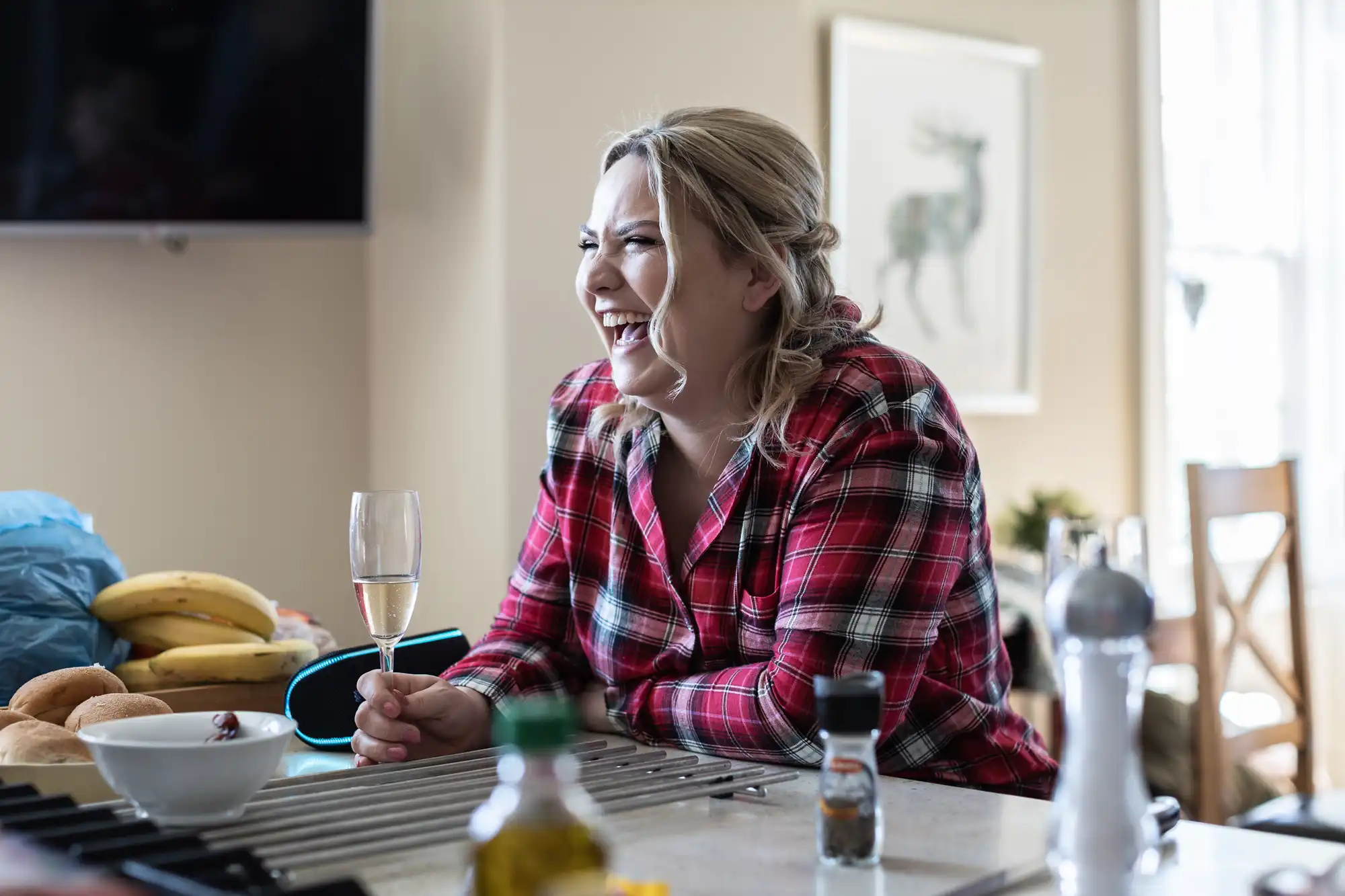 A person in a plaid shirt smiles and holds a champagne glass while seated at a kitchen counter with various items, including bananas, bread rolls, and a pepper grinder, in view.