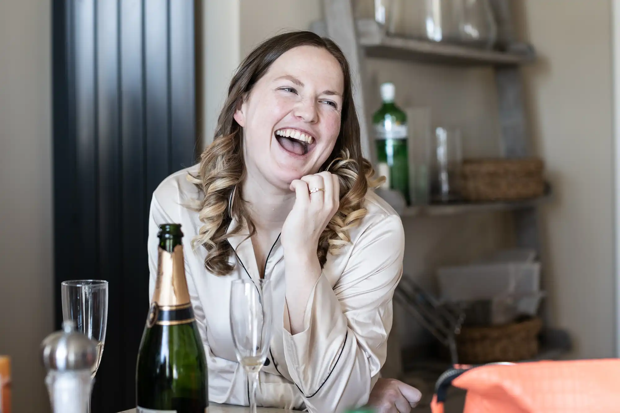 A woman dressed in light clothing is sitting at a table, smiling and laughing. There are champagne glasses and a bottle on the table, with a shelf in the background holding various items.