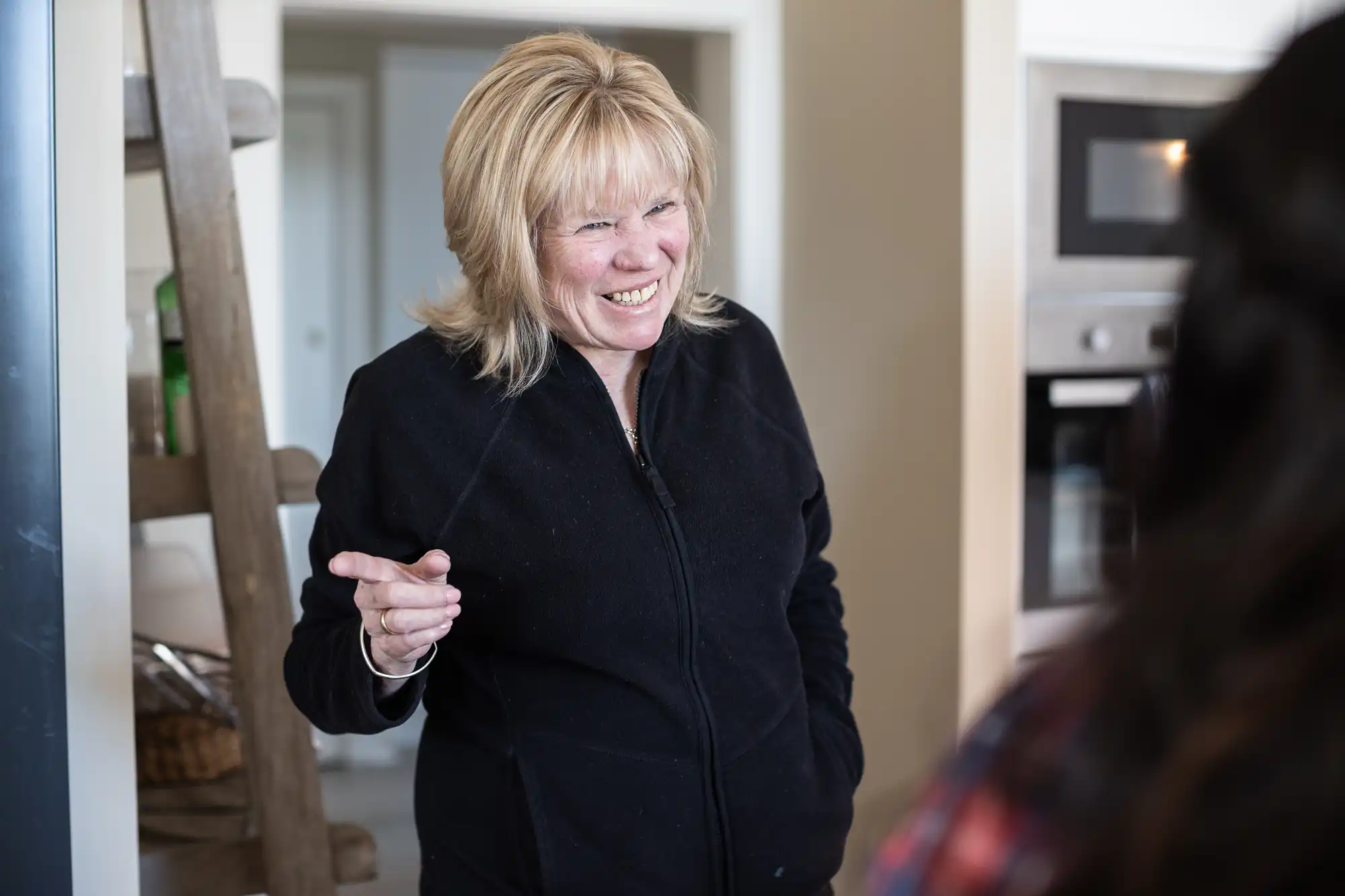 A woman with light hair, wearing a black jacket, smiles and gestures with her hand while standing in a kitchen with a ladder and an oven in the background.