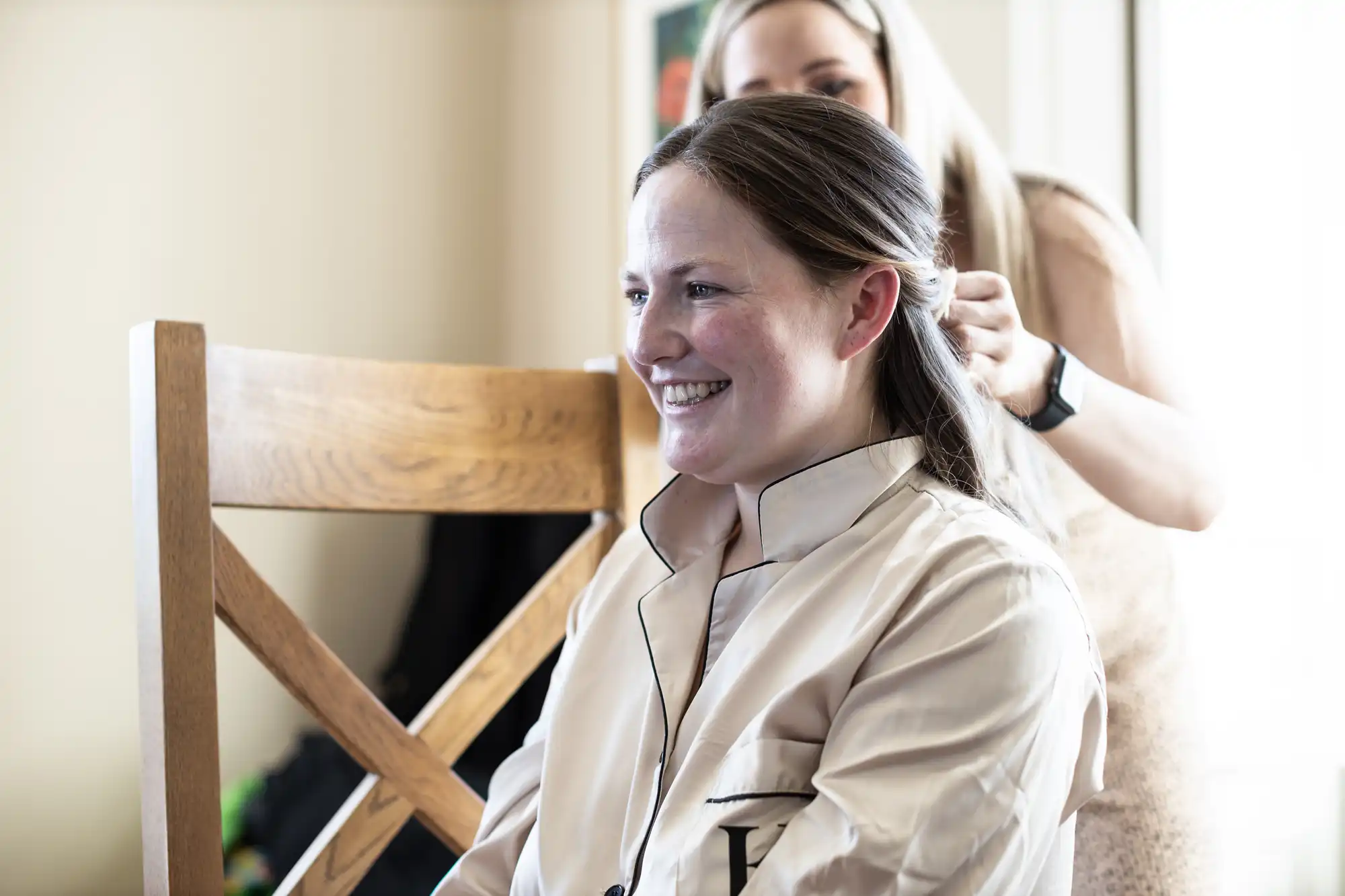 A woman with a smile, seated on a wooden chair, is having her hair styled by another woman standing behind her.