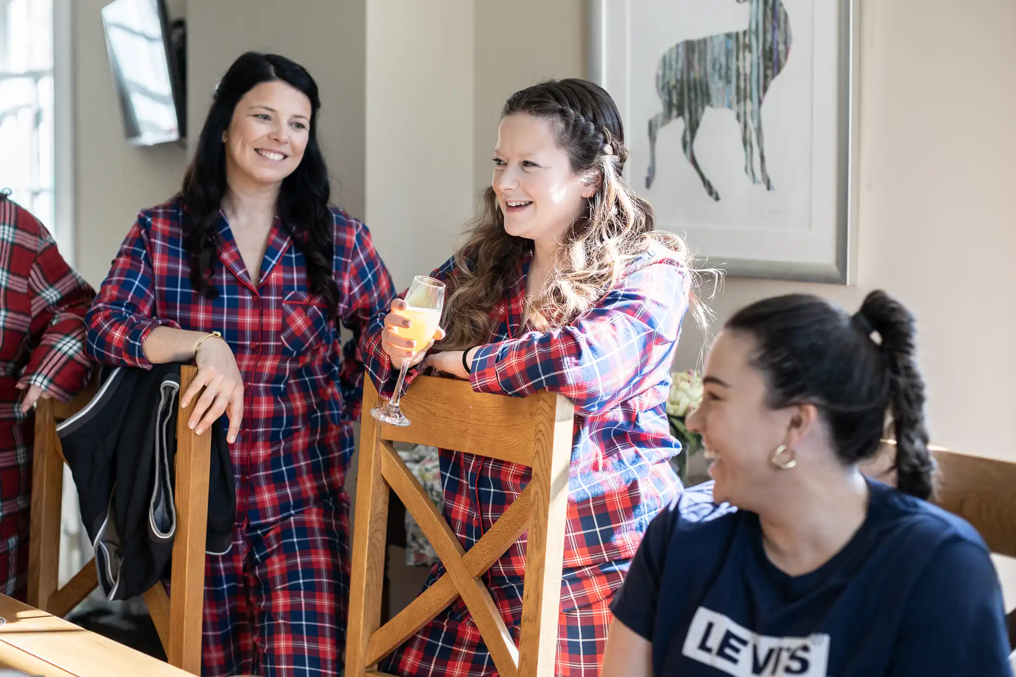 Three women are having a conversation in a well-lit room. Two of them are in plaid pajamas, with one holding a drink, and the third woman is smiling while wearing a Levi's t-shirt.